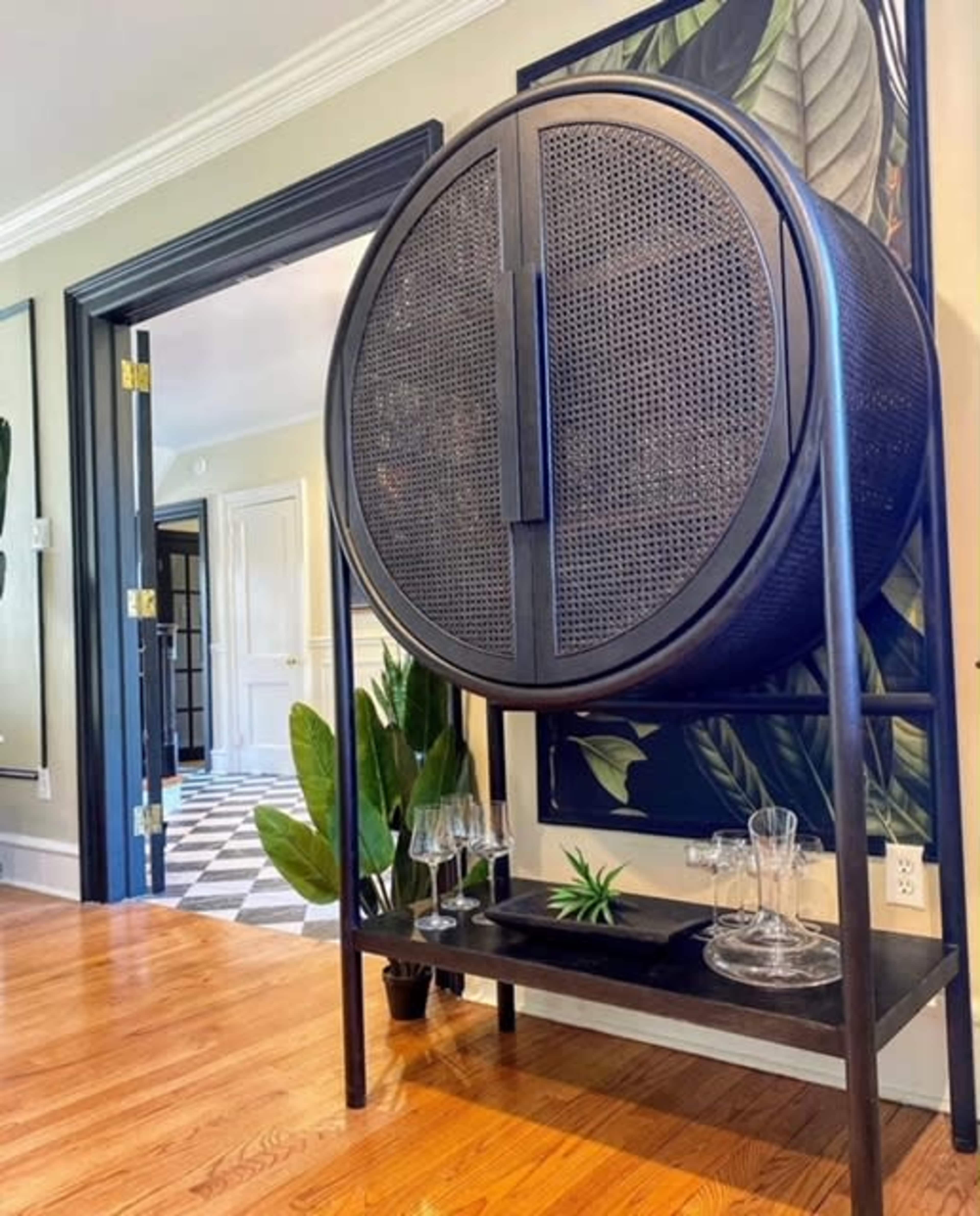 A round, black woven cabinet stands on a wooden floor, accompanied by several glassware items and a small plant, with a patterned wall and open doorways in the background.