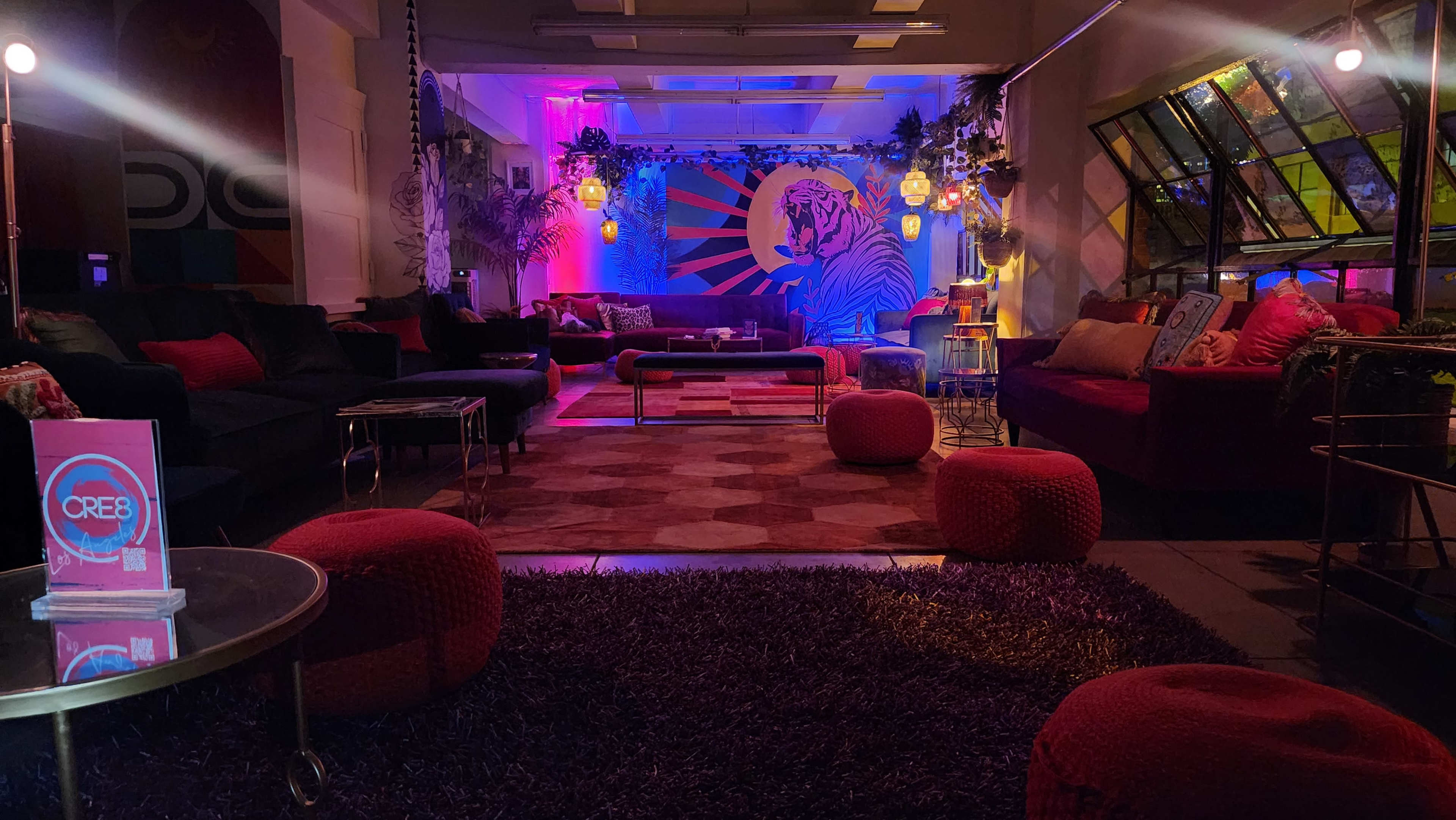 The image shows a vibrant lounge area featuring plush seating, colorful lighting, and a large mural of a tiger on the wall.