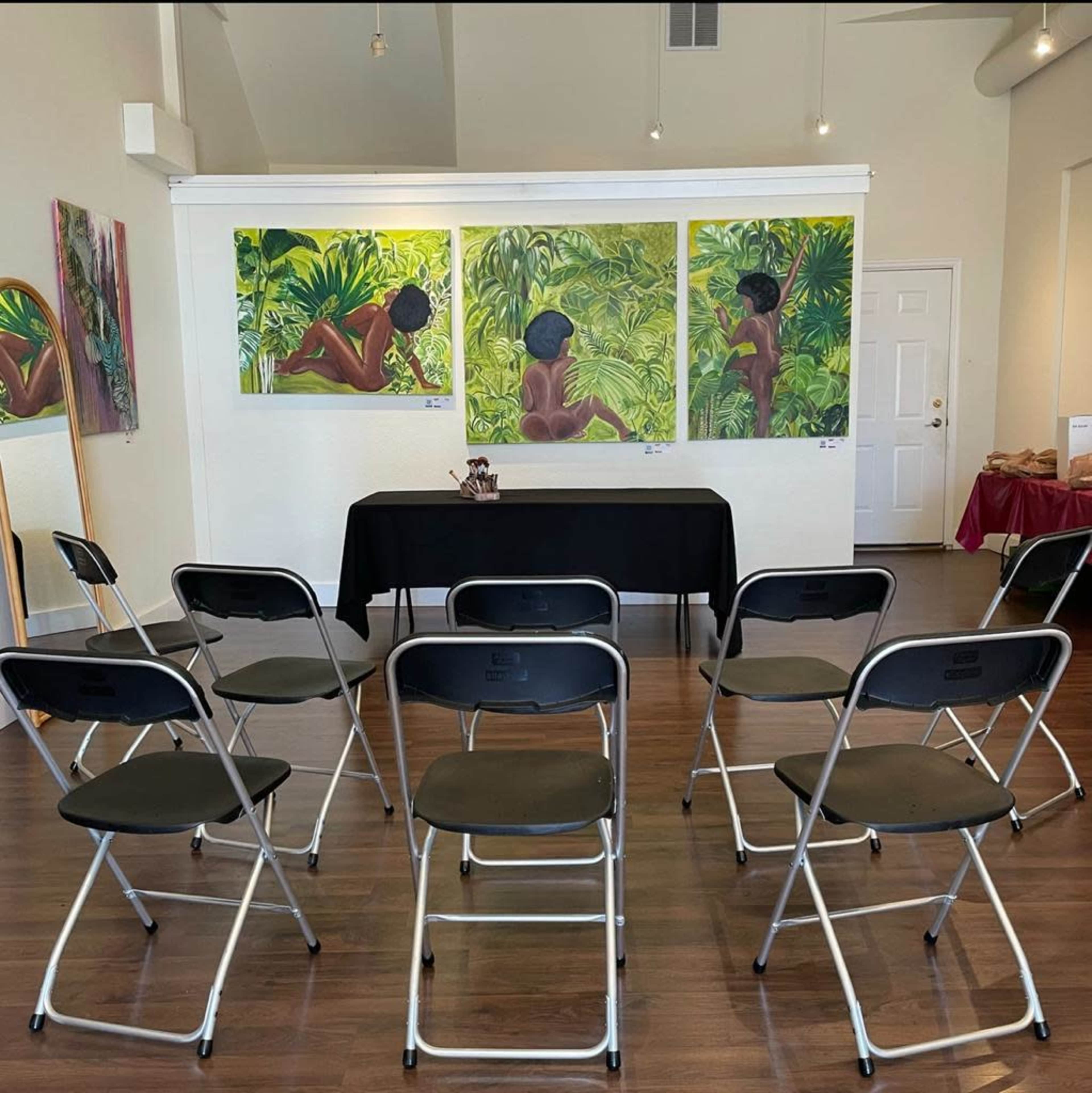 An art gallery featuring three large paintings of figures in a tropical setting, with several folding chairs arranged in front for viewers.