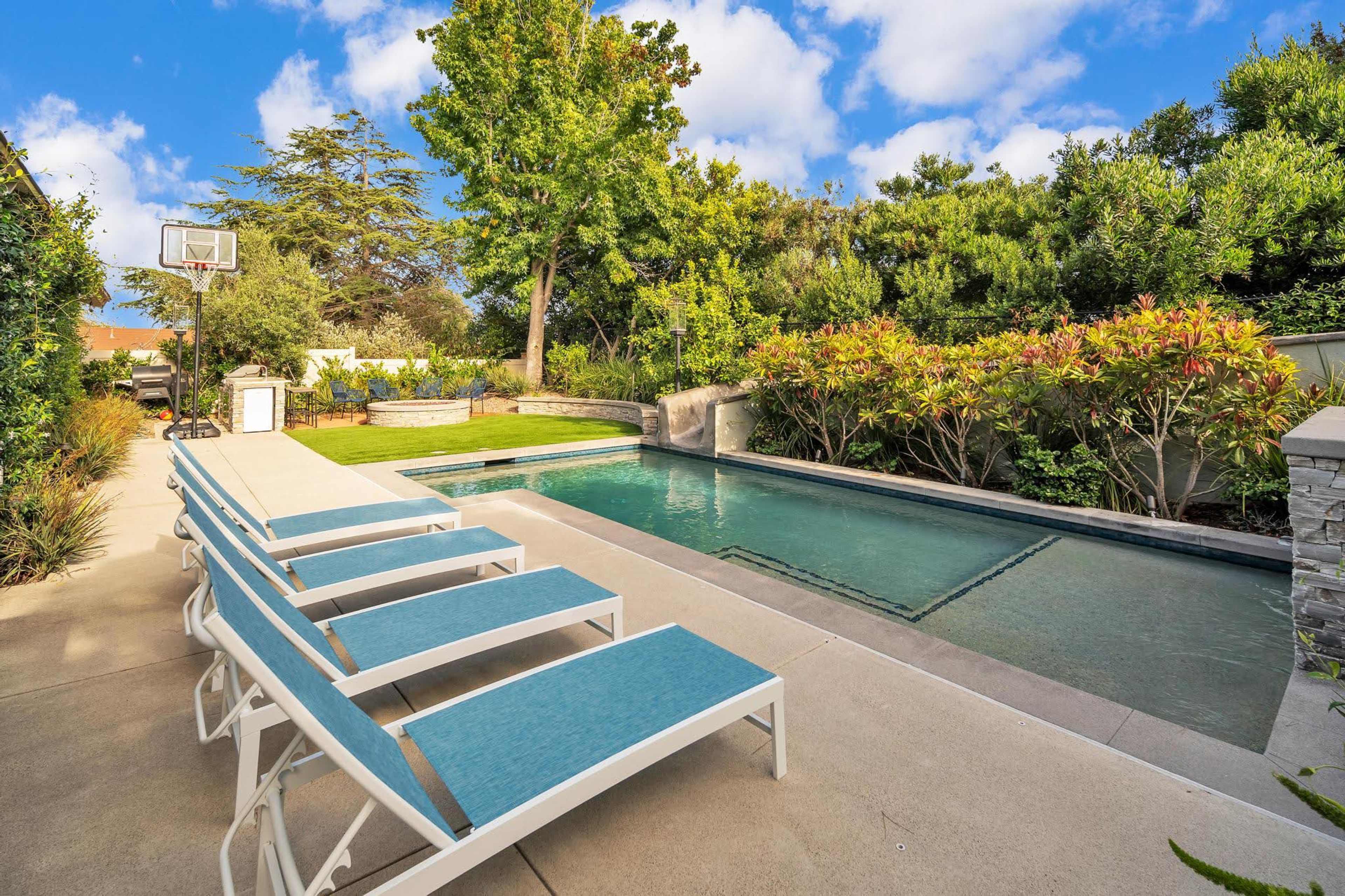 A backyard with a swimming pool surrounded by lounge chairs, landscaped greenery, and a basketball hoop in the background.