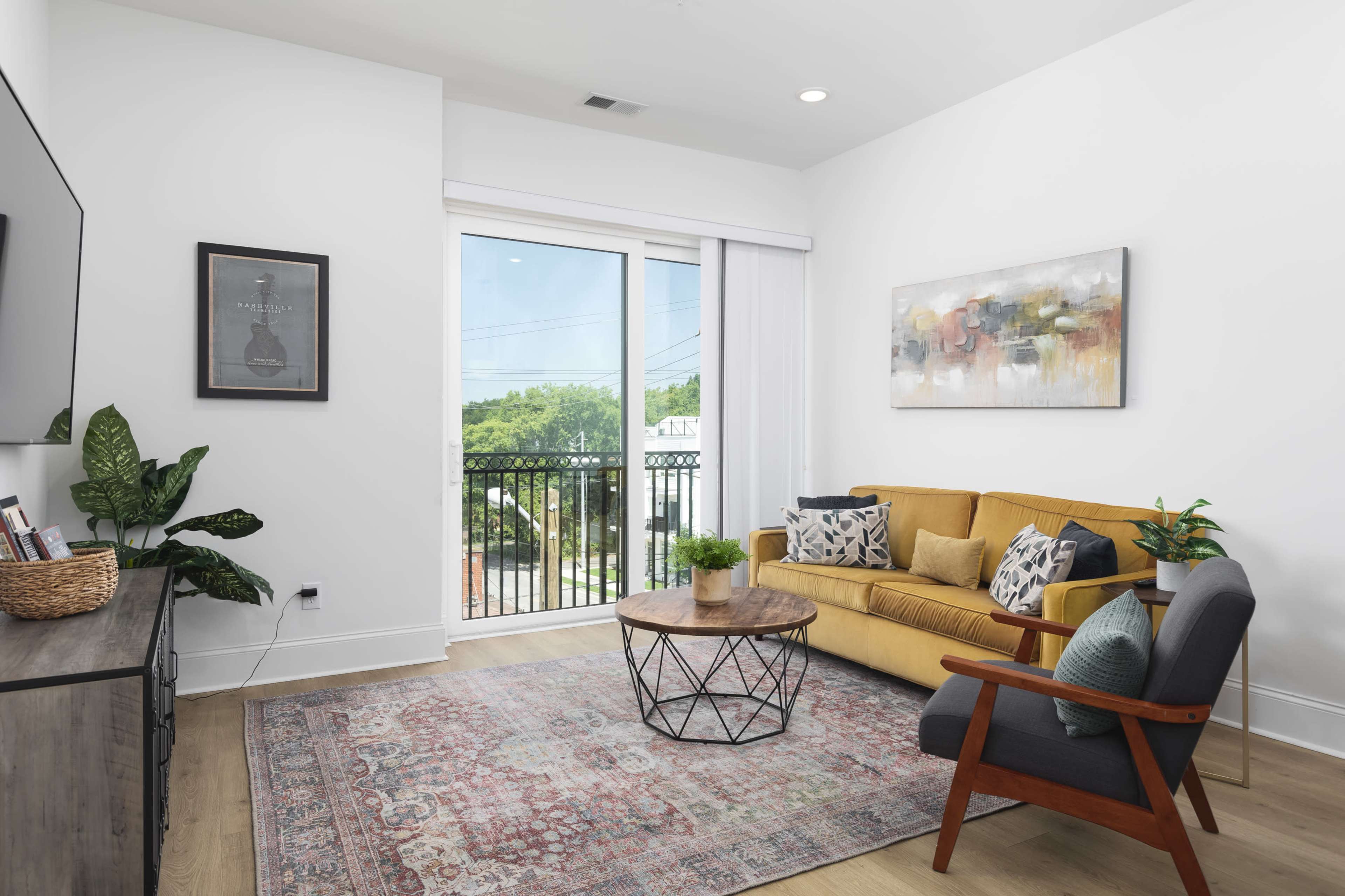 The image depicts a modern living room featuring a yellow sofa, a coffee table with a geometric base, and a view of the outdoors through large sliding glass doors.