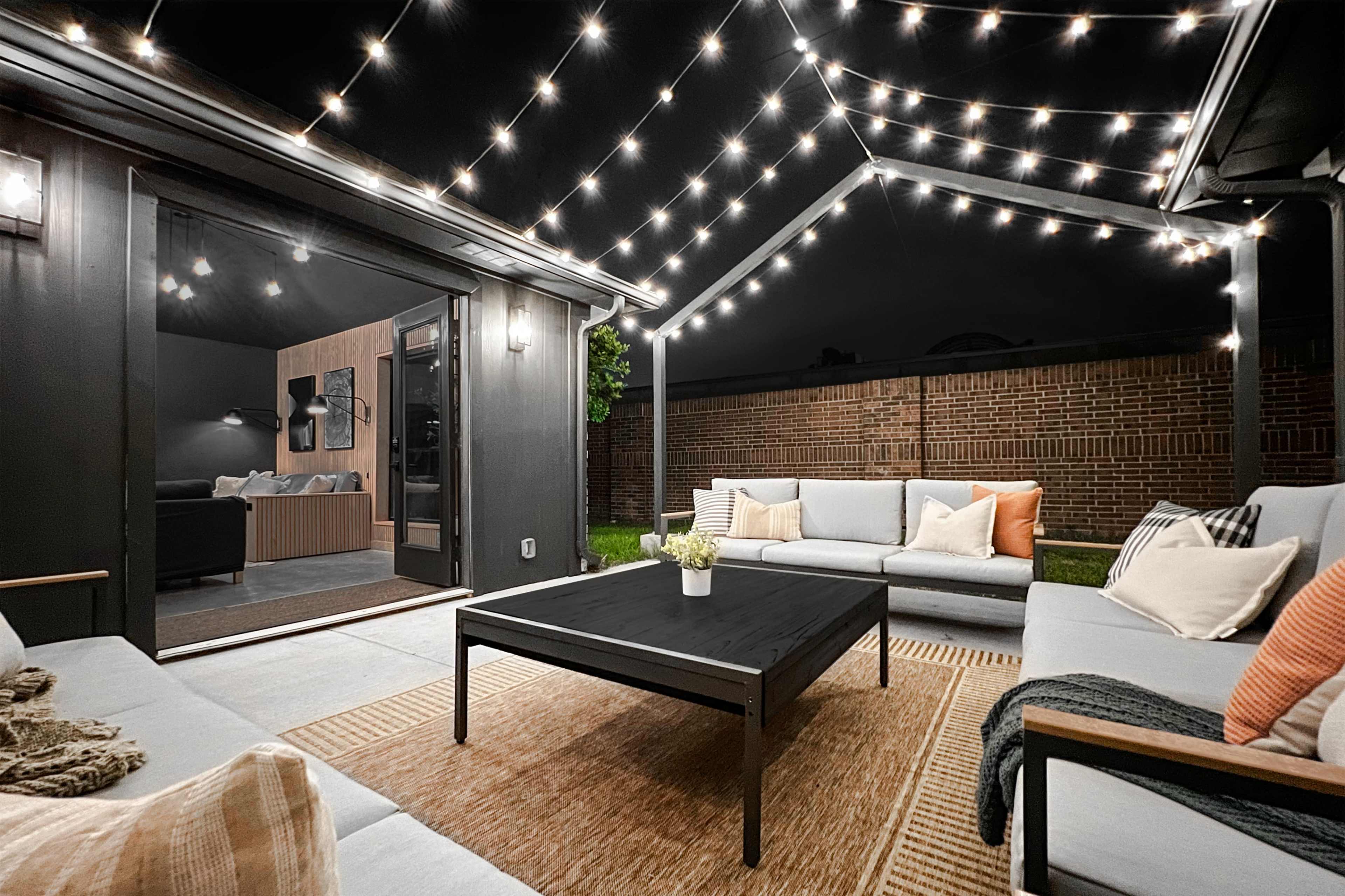A patio area features a seating arrangement with light-colored couches and a dark coffee table, illuminated by string lights overhead.