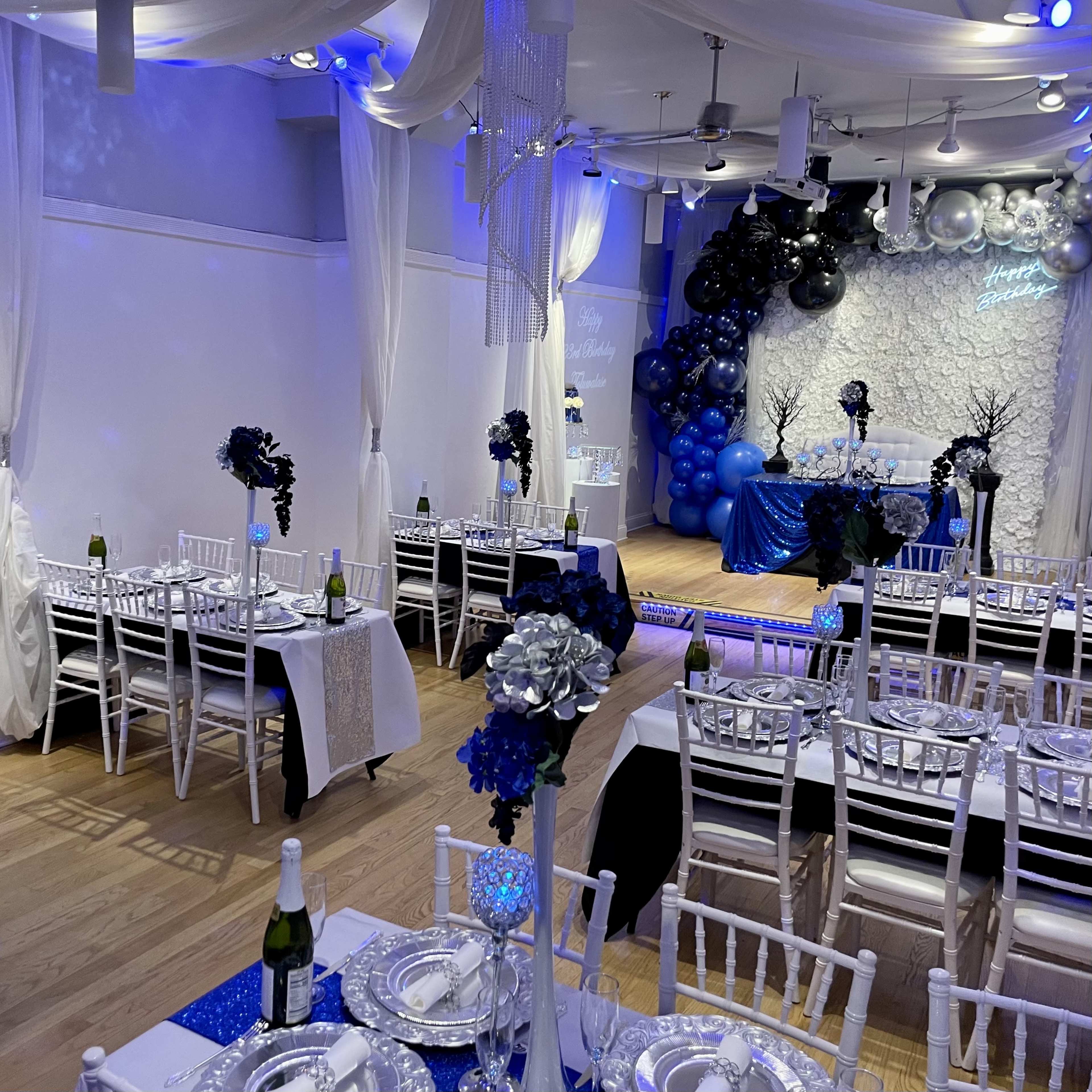A decorated event space features white and black tables with blue accents, adorned with floral centerpieces and shimmering tableware, set for a celebration.