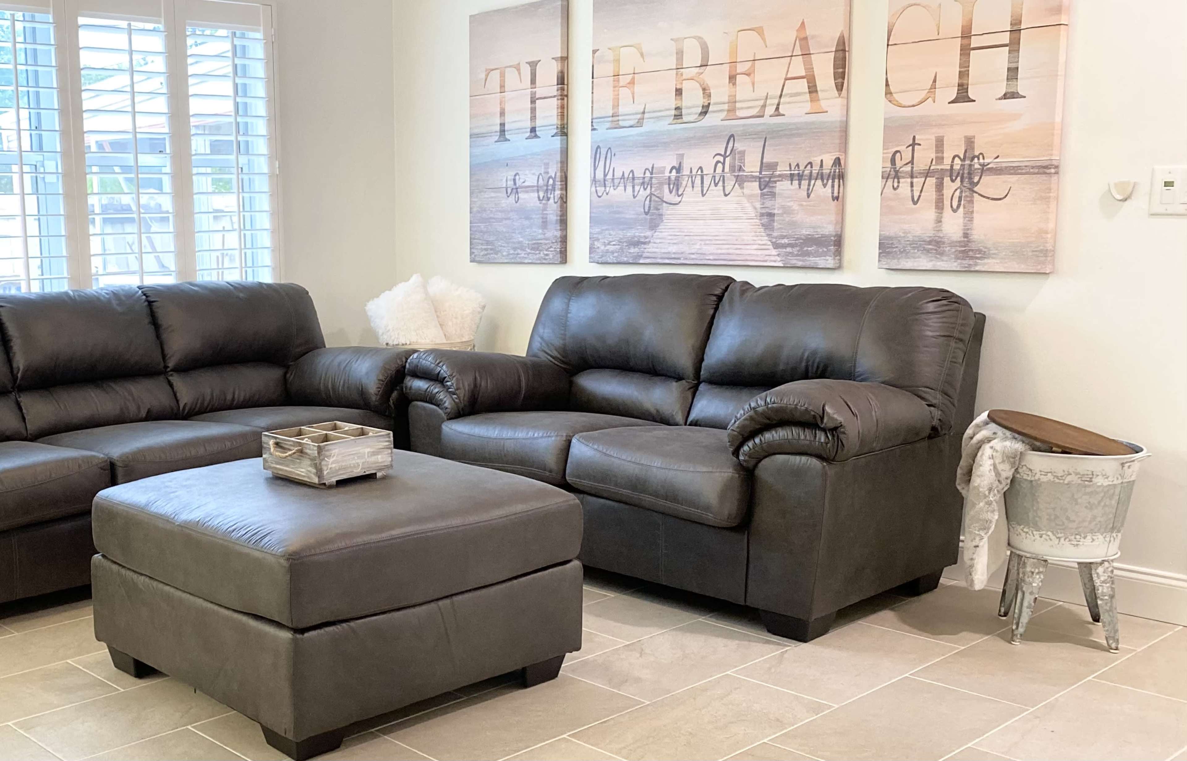 A living room features a gray leather sofa and armchair, accompanied by a matching ottoman and decorative wall art that reads "THE BEACH."