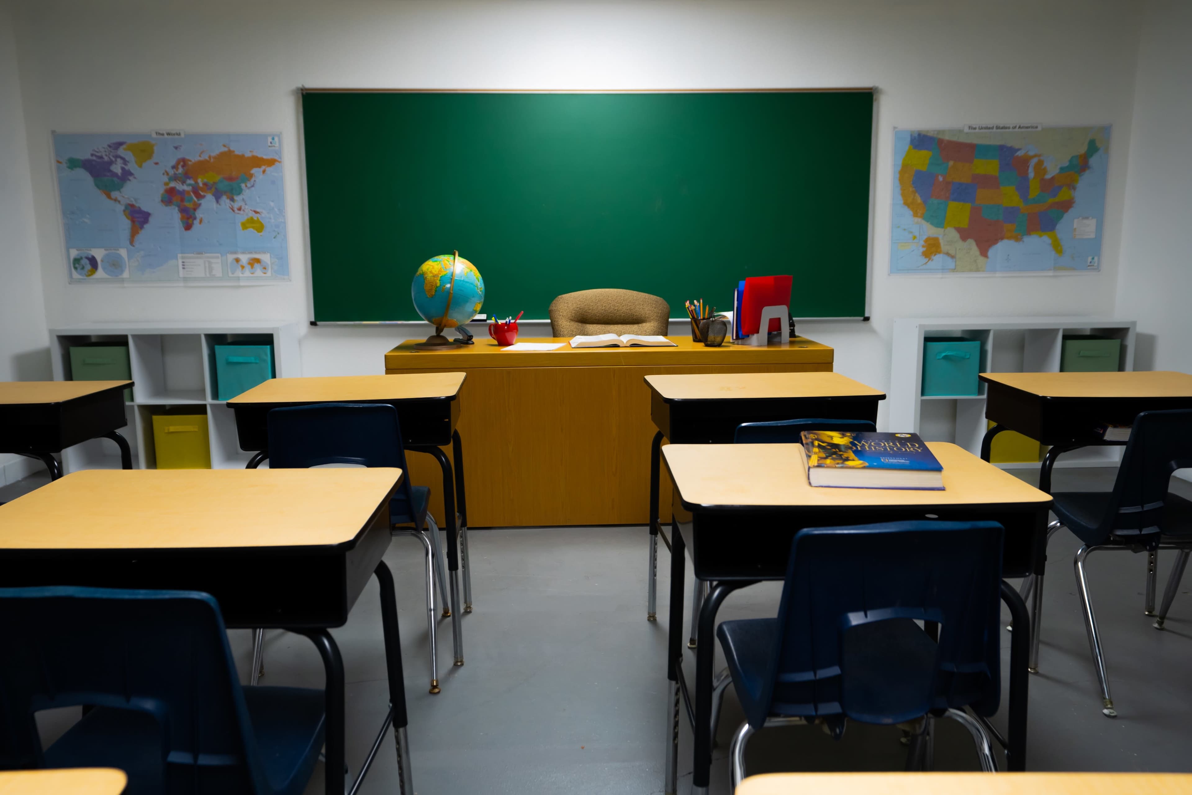 A classroom is arranged with empty desks facing a teacher's desk, which is positioned in front of a green chalkboard and surrounded by maps on the walls.