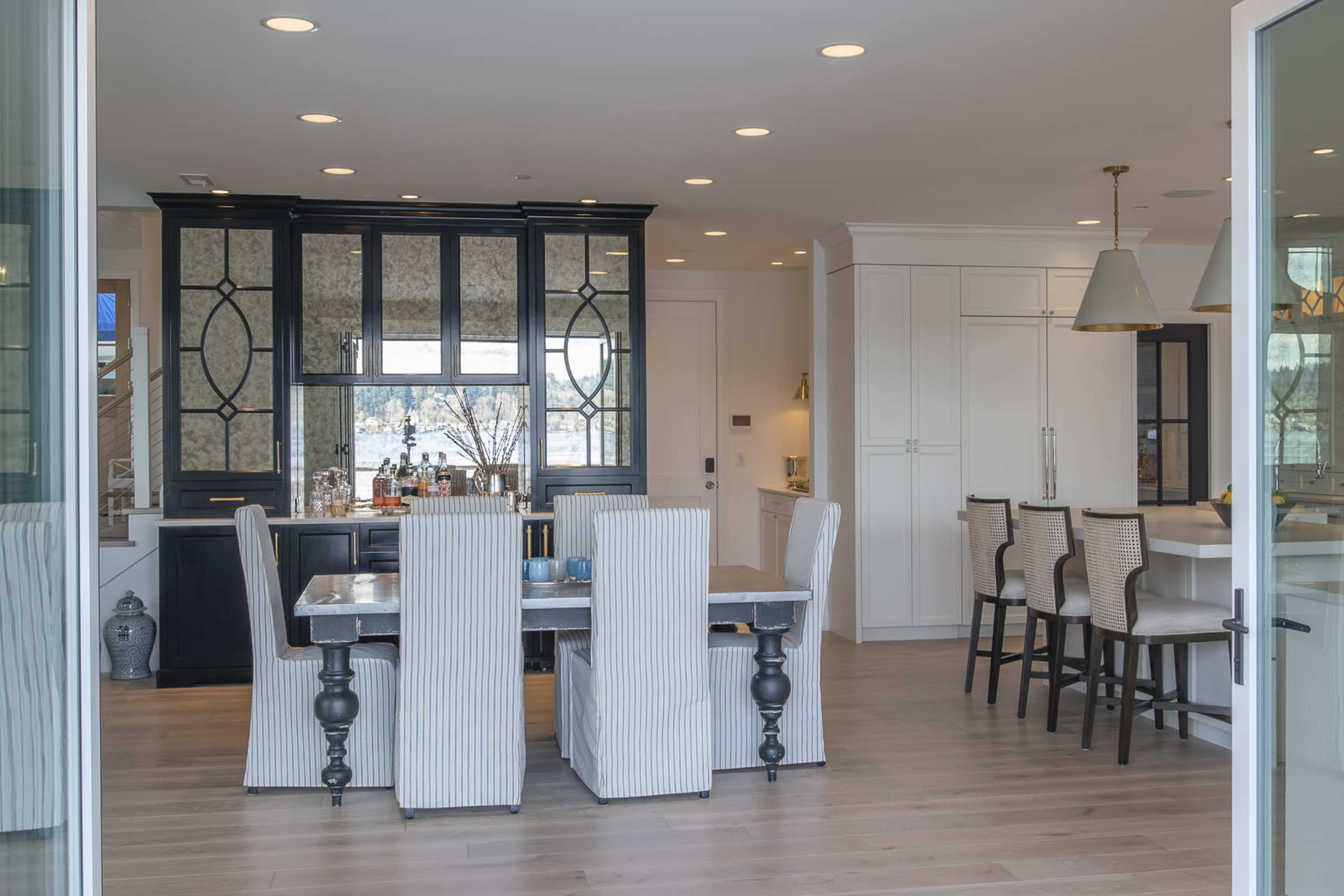 The image shows a modern kitchen and dining area featuring a dark cabinetry unit, a large dining table with striped chairs, and open access to a well-lit cooking space.