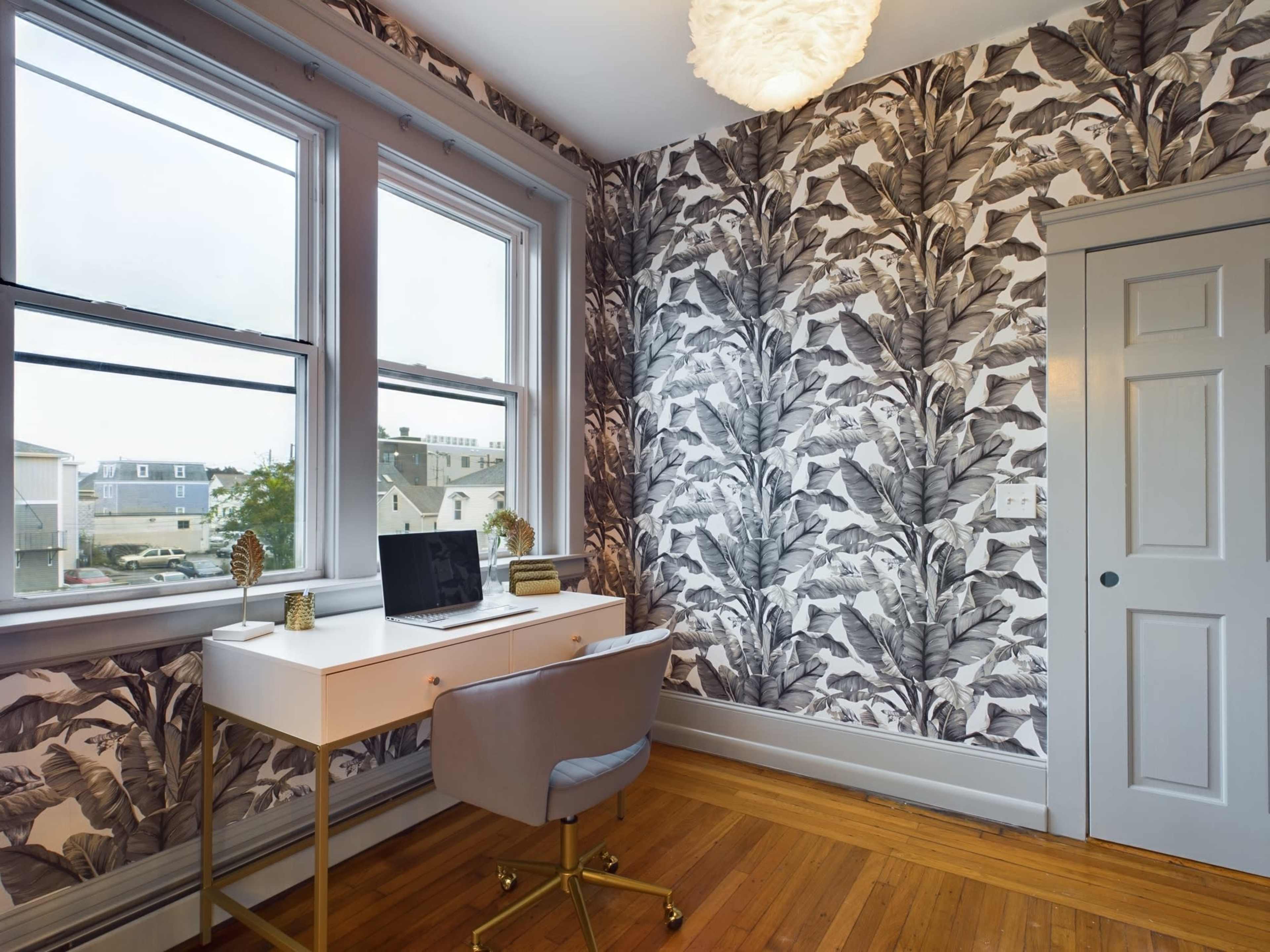 A home office features a white desk with a laptop in front of patterned wallpaper depicting large leaves, and a gray chair on a wooden floor next to a door.