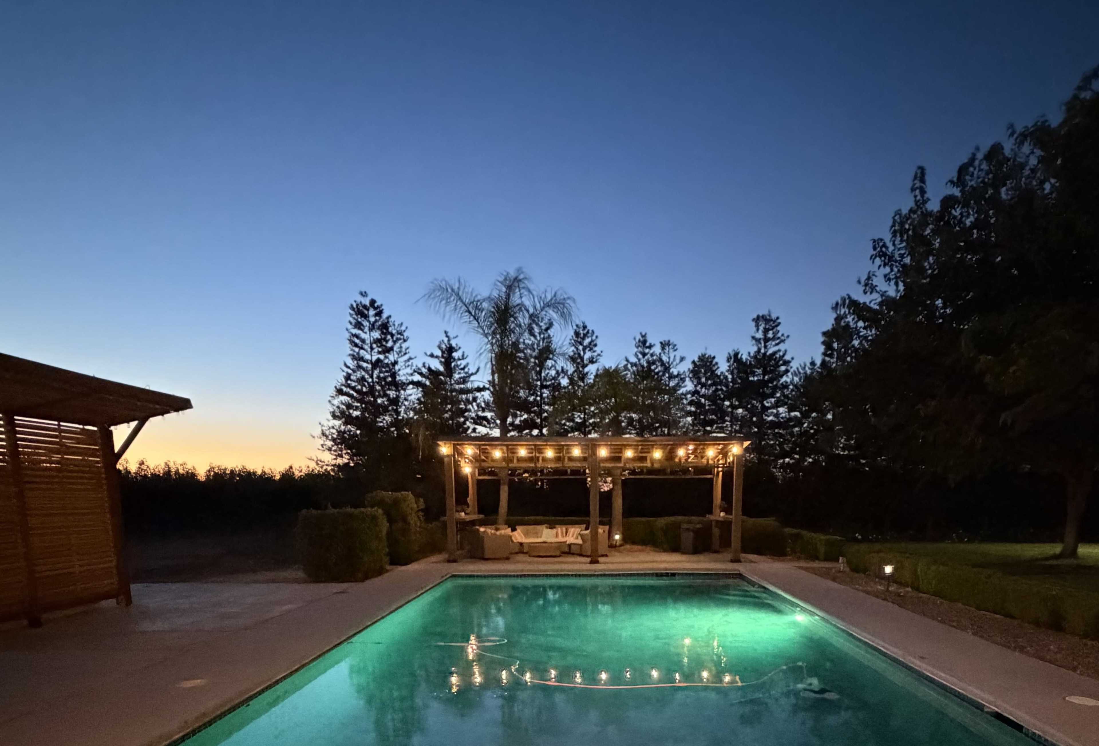 A well-lit pergola with seating overlooks a clear swimming pool at dusk, surrounded by trees and a gradient sky.