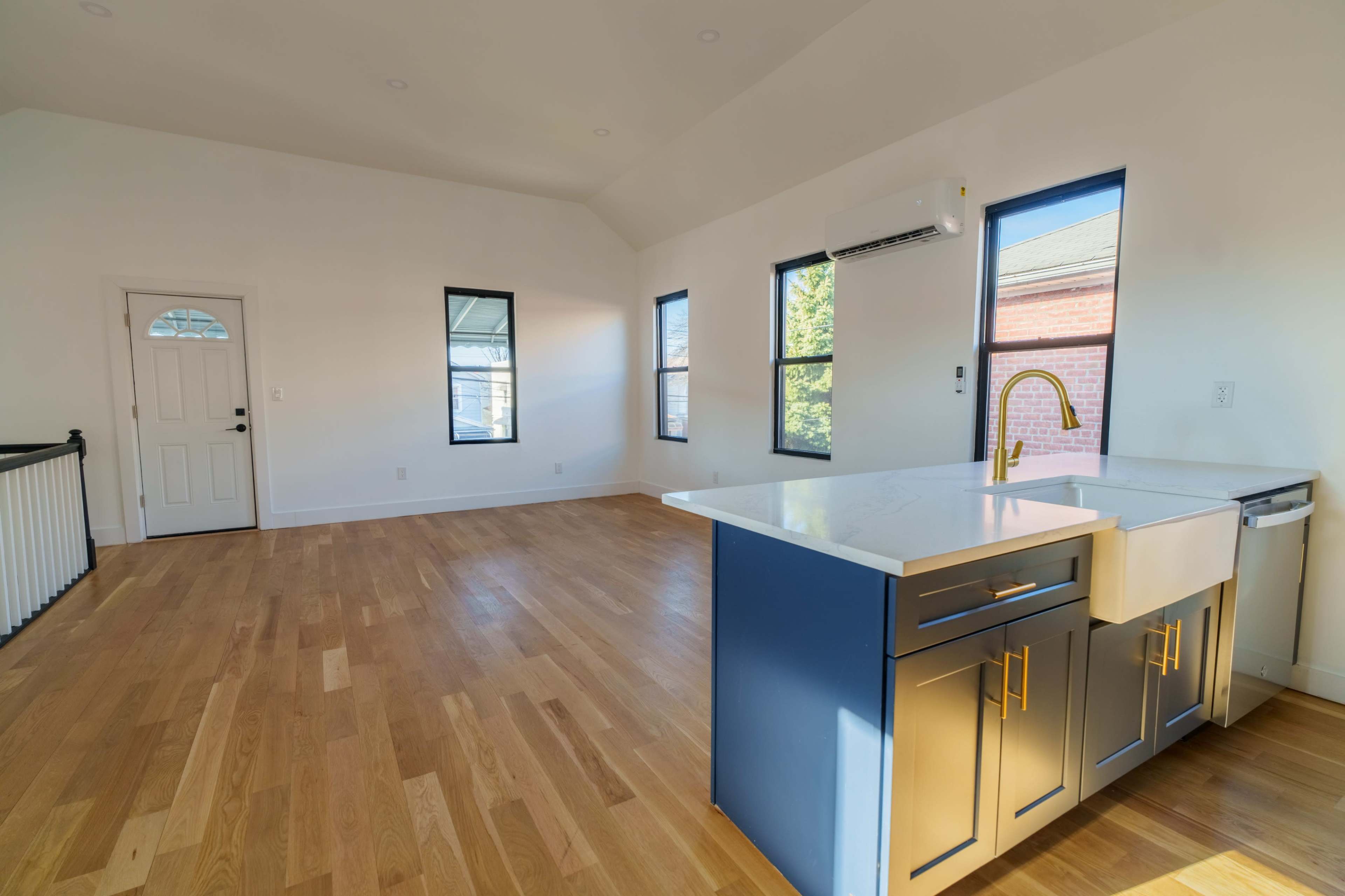 The image shows a spacious, well-lit room with large windows, hardwood flooring, and a kitchen area featuring a sink and cabinetry.