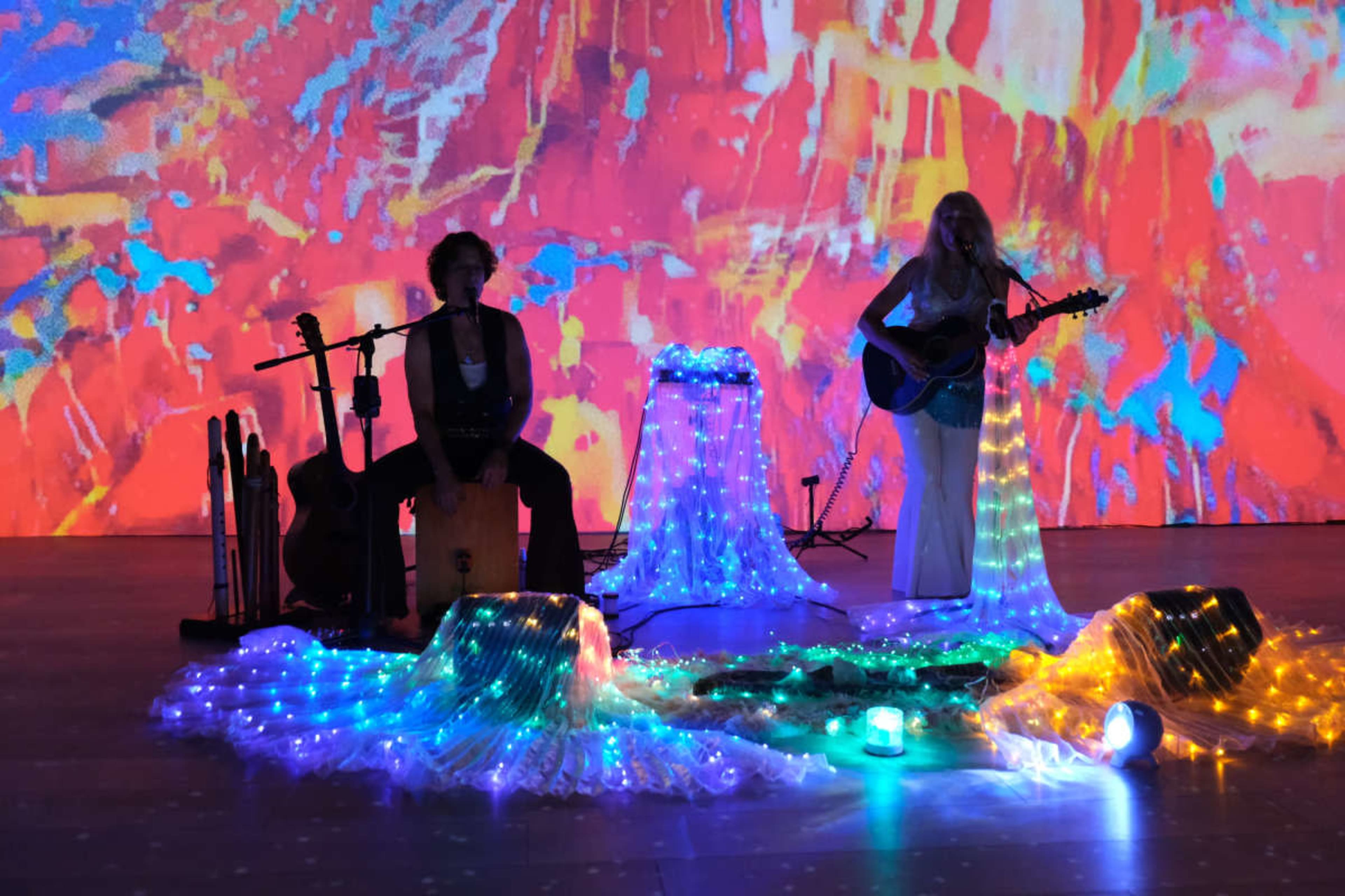 A musical duo performs on stage with colorful, illuminated backdrops and decorative light strands.