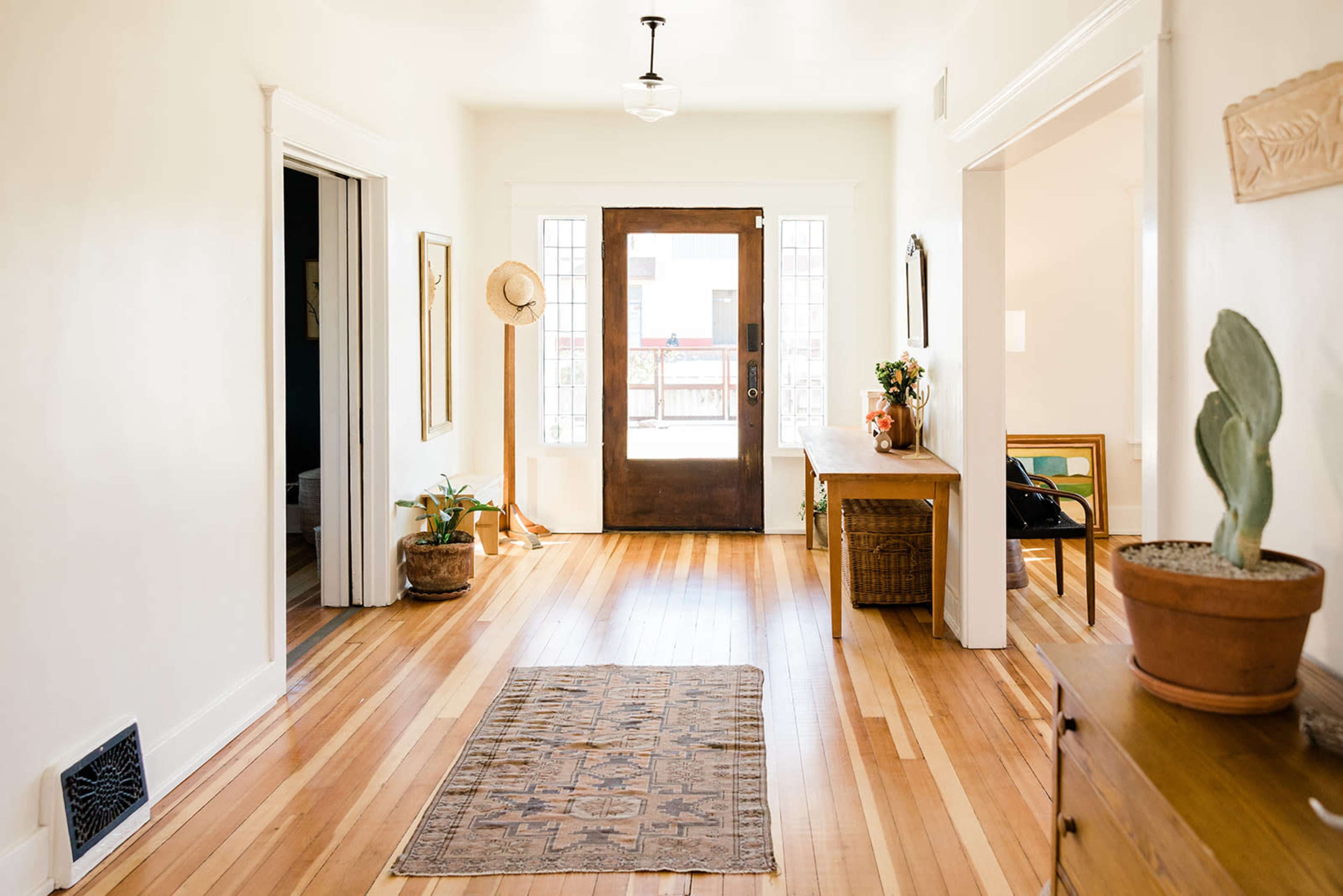 A bright hallway features wooden floors, a front door with glass panels, and decor including a vintage rug, a potted plant, and wall art.