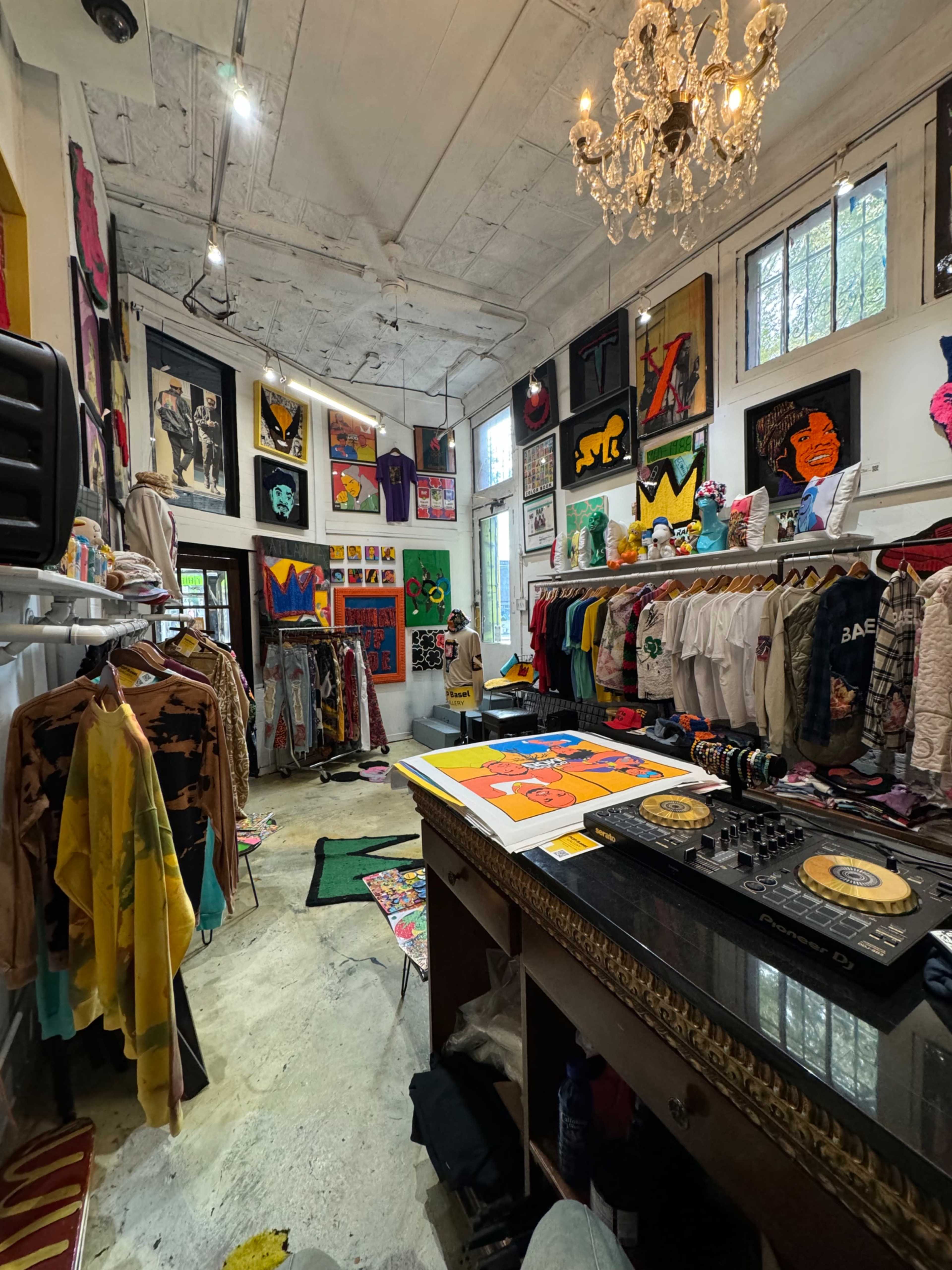 A colorful boutique features clothing racks filled with vibrant apparel and artwork adorning the walls, along with a DJ setup in the foreground.