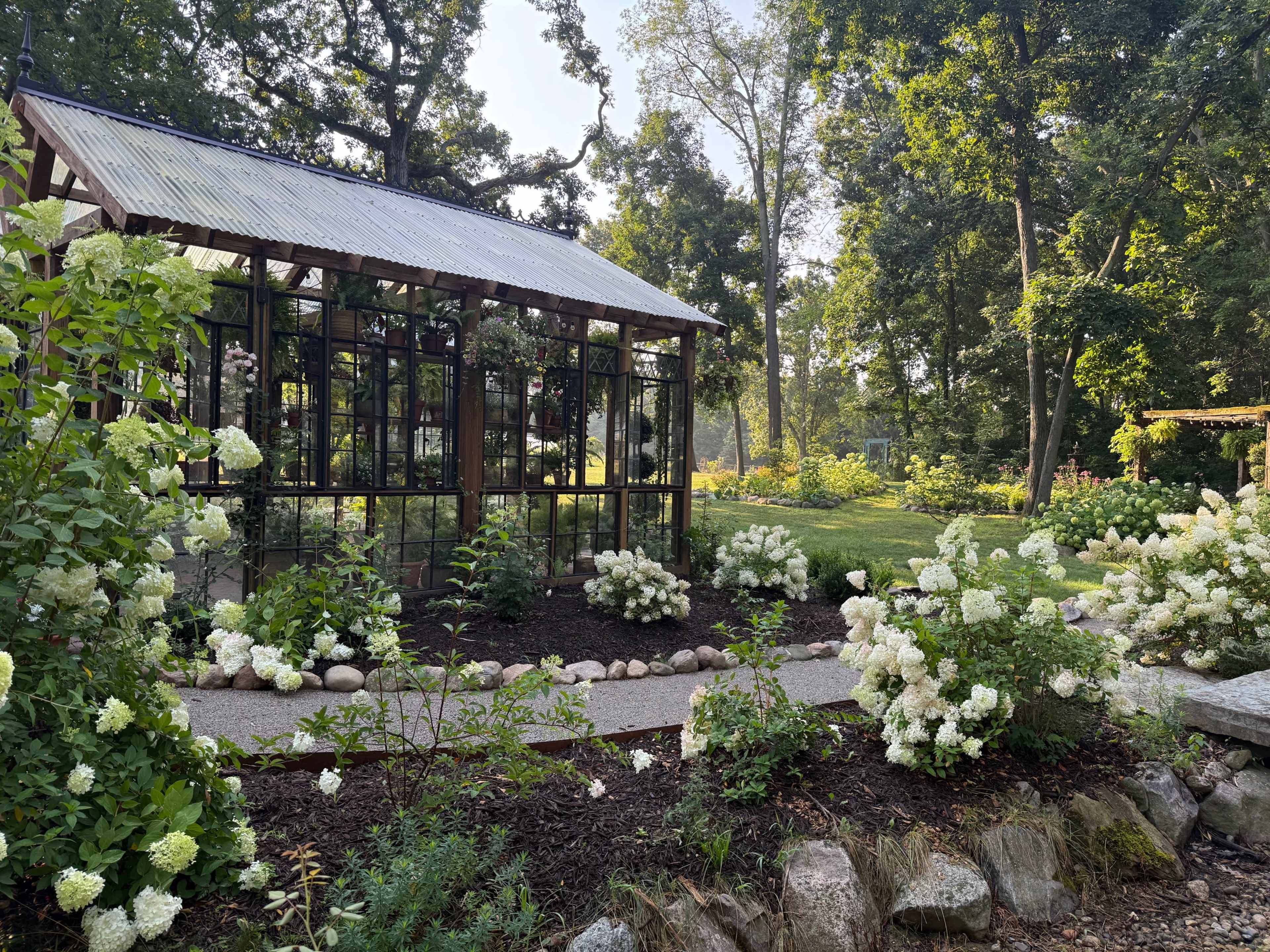 A greenhouse made of glass and wood stands among lush greenery and flowering bushes in a sunlit garden.