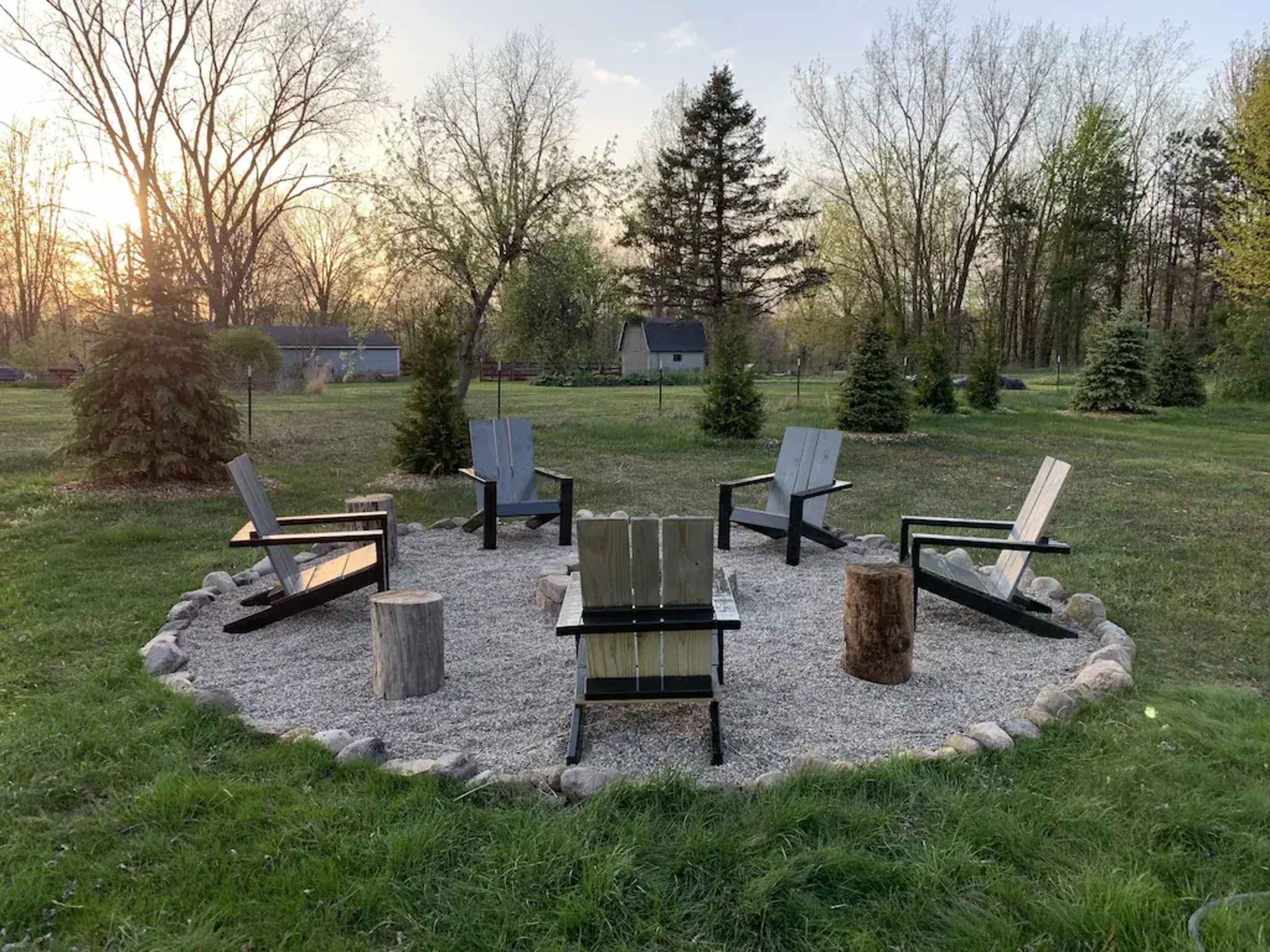 A circular arrangement of six wooden chairs around a gravel area, surrounded by grass and trees, with a sunset in the background.