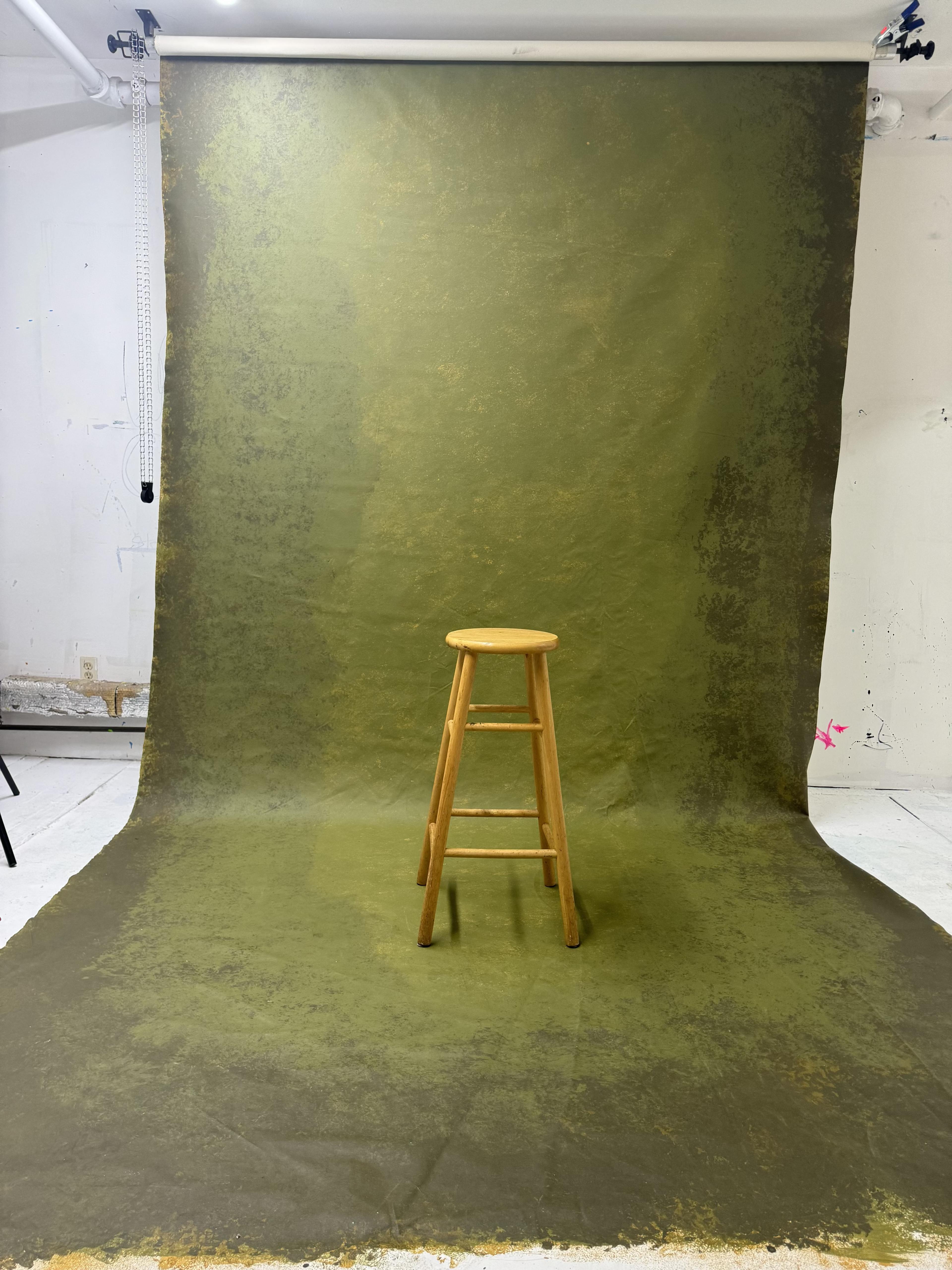 A wooden stool is positioned in front of an olive green backdrop.