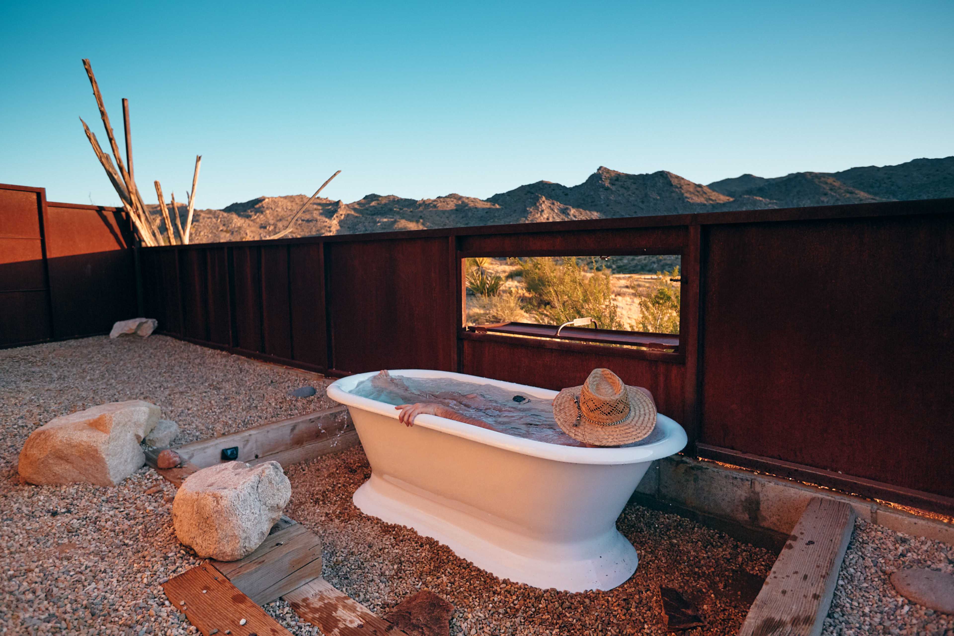 A white bathtub sits on a gravel surface, surrounded by rocks, with a straw hat resting on the edge, framed by a backdrop of mountains under a blue sky.