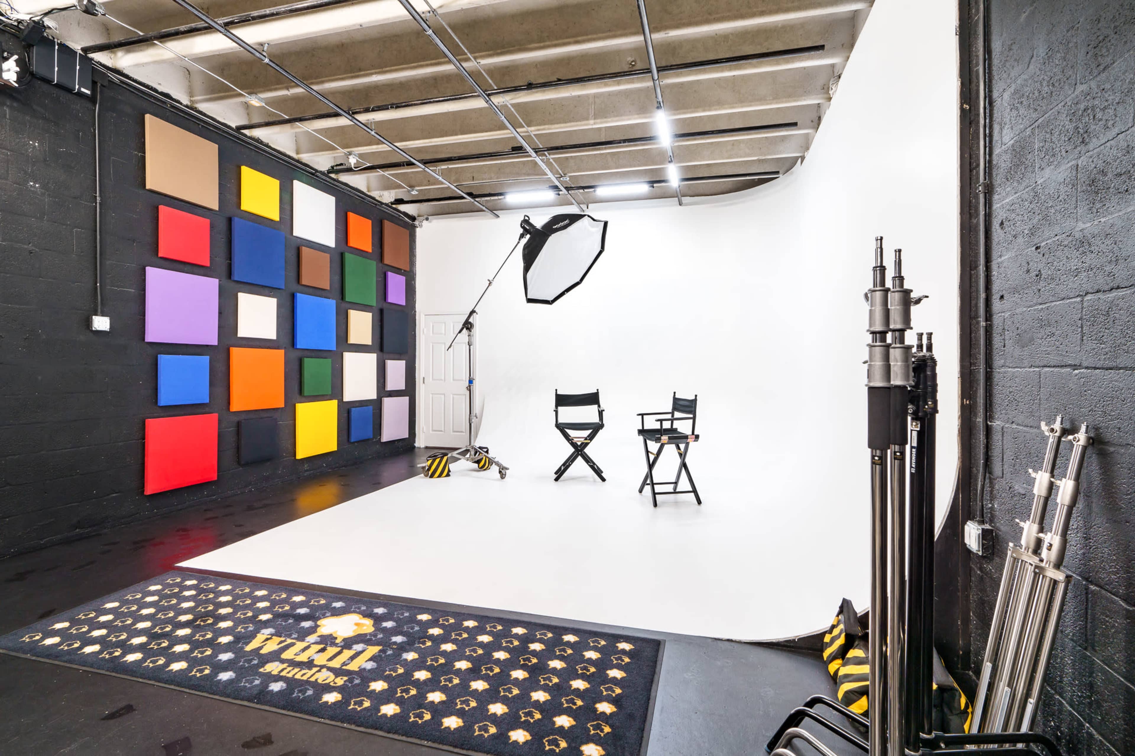 A photography studio with a white backdrop, two director's chairs, and a colorful wall display of squares on one side.