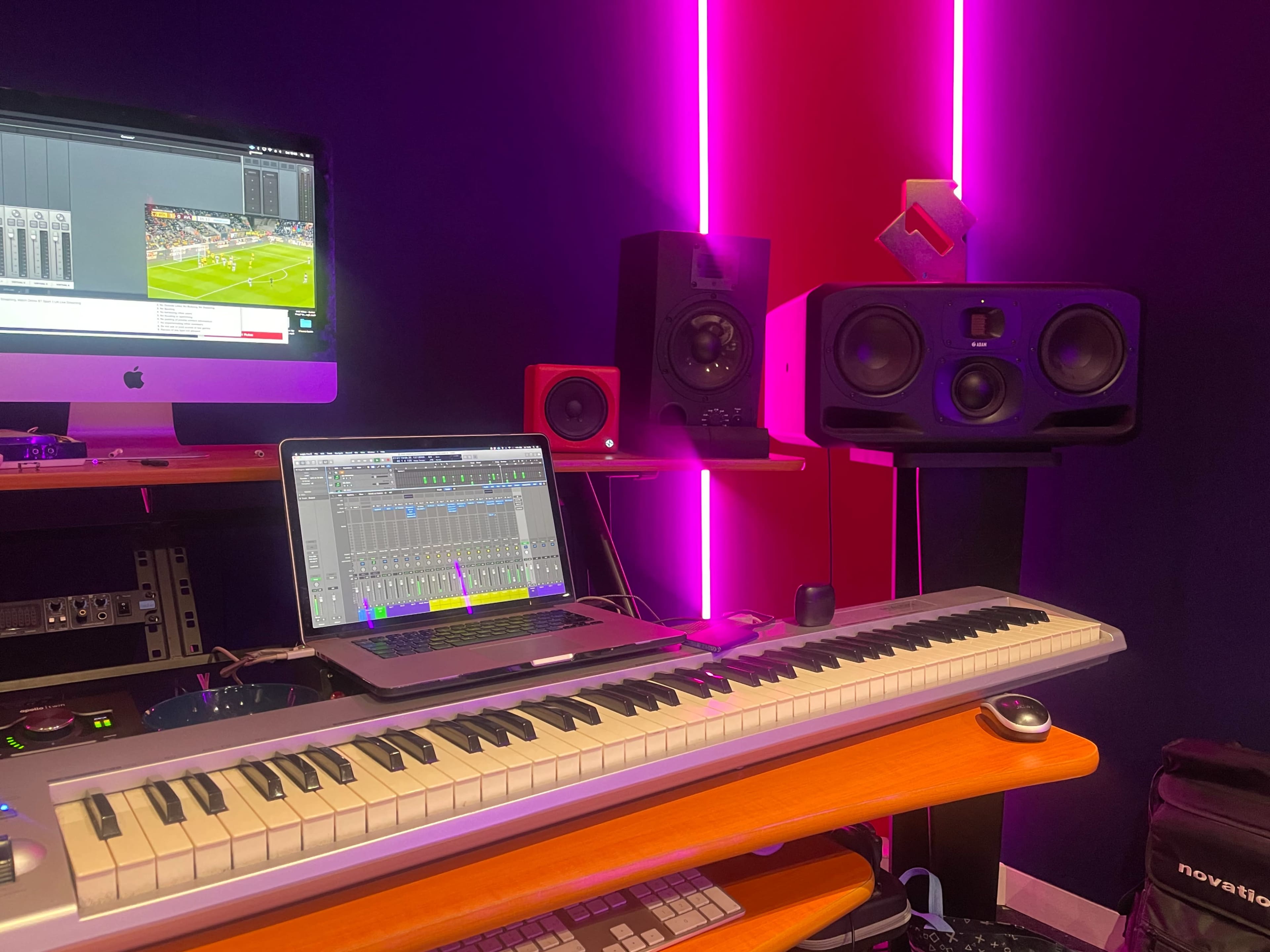 A music production workspace features a MIDI keyboard, a computer displaying audio software, and large studio monitors against a backdrop of vibrant neon lighting.