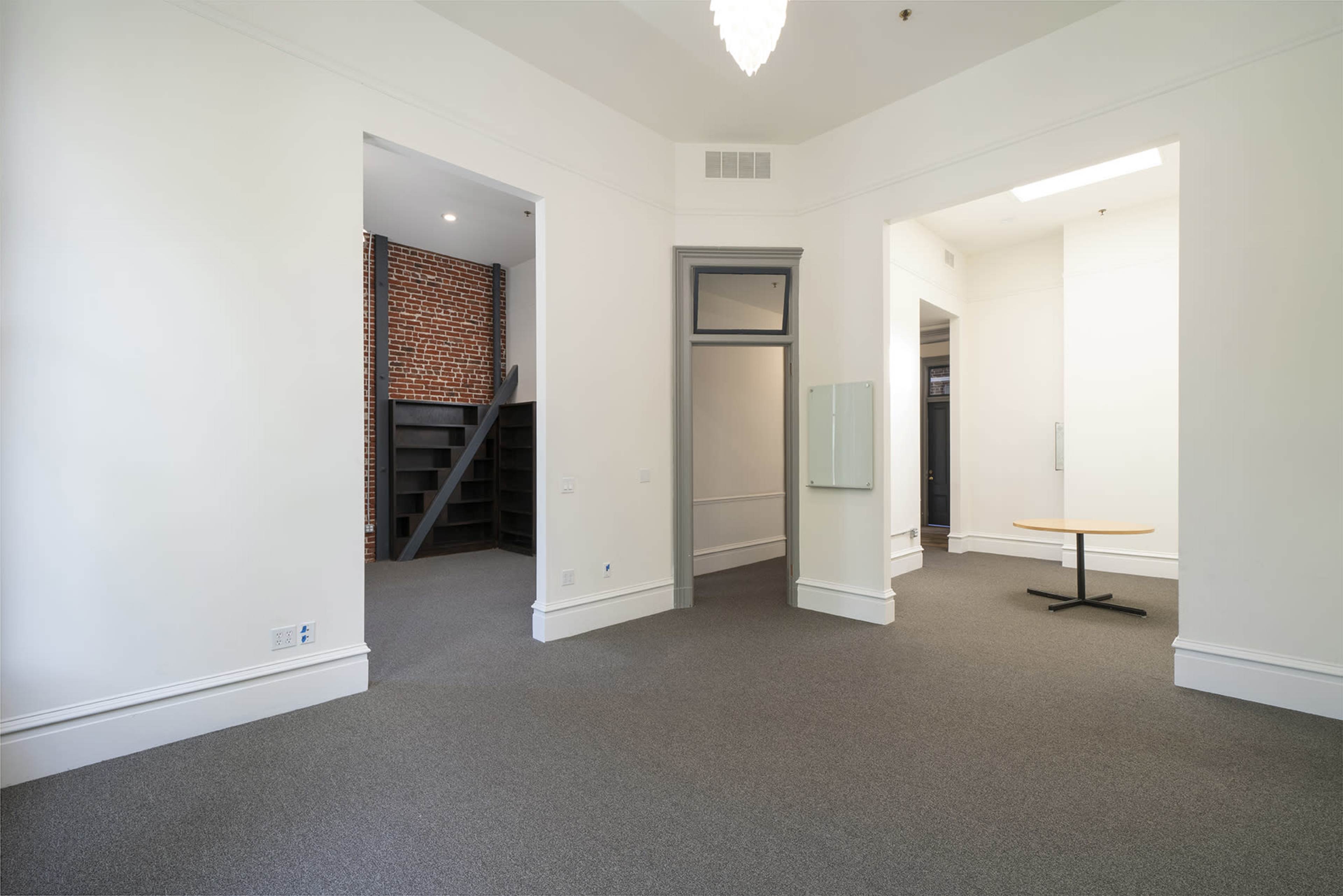 A spacious, minimally furnished room with light-colored walls, a carpeted floor, and a staircase leading to an upper level in the background.