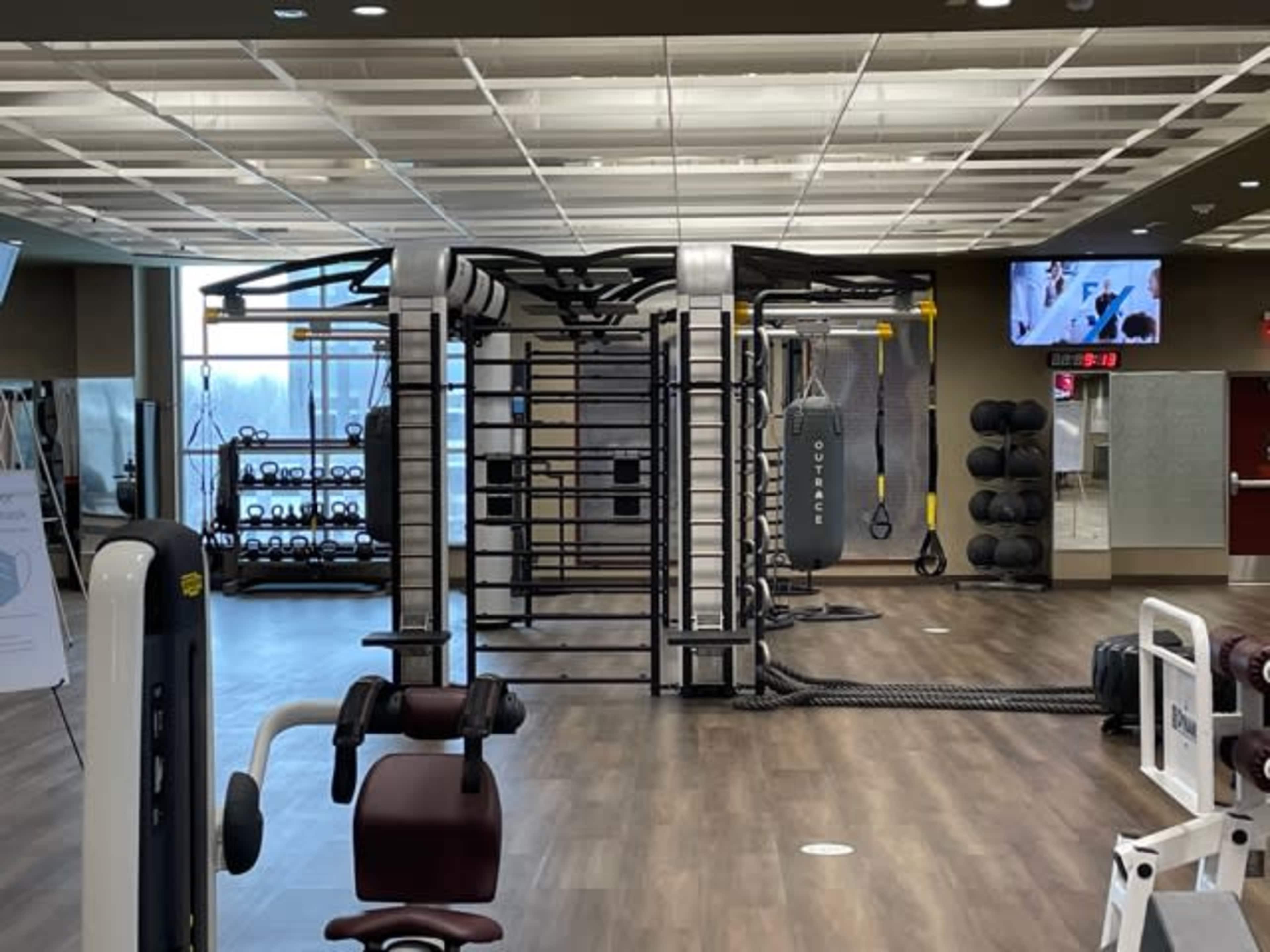 The image shows a modern gym interior with various exercise equipment, including weight machines and a suspension training rig.