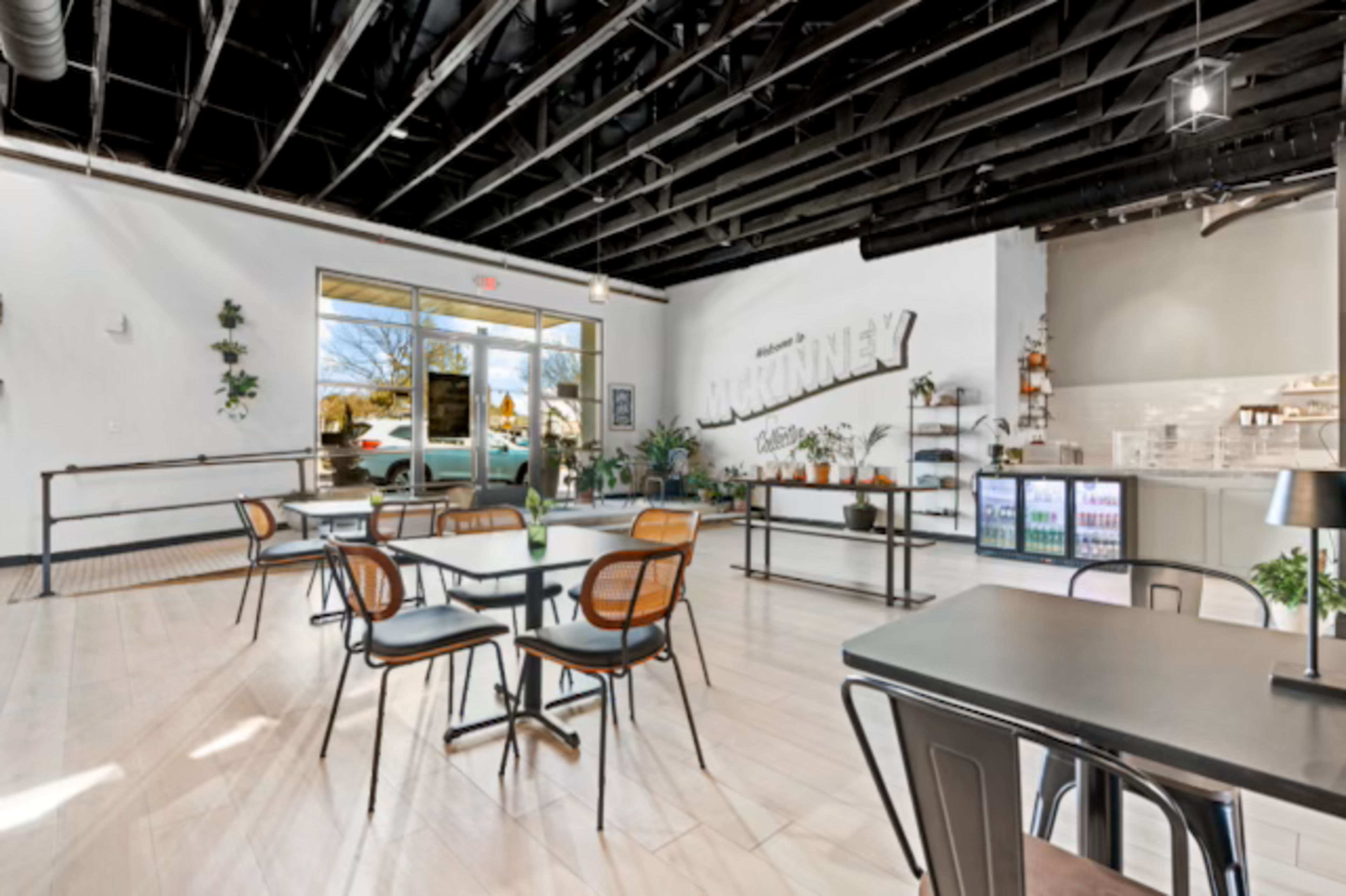 The image shows a modern café interior with tables and chairs, large windows, and plants decorating the space.