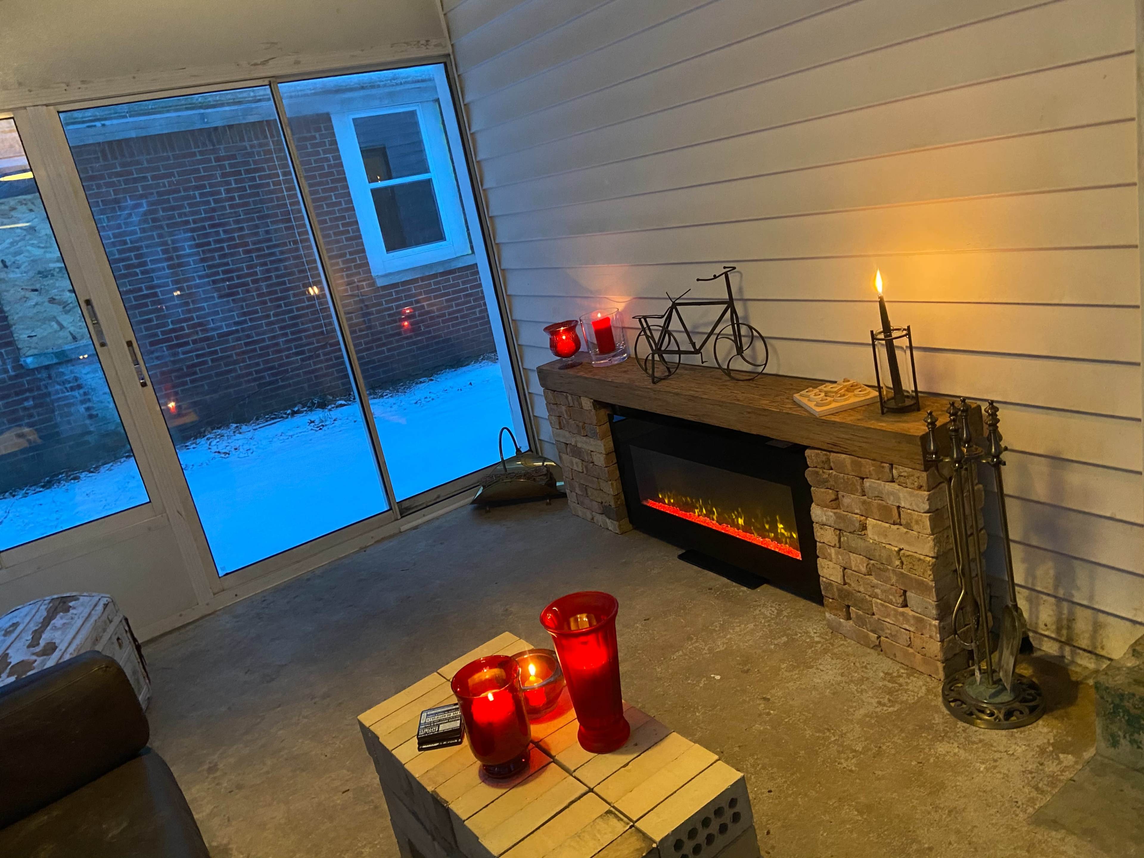 A cozy indoor space with a wooden mantel, candles in red glass holders, an electric fireplace, and a view of snow outside through large glass doors.