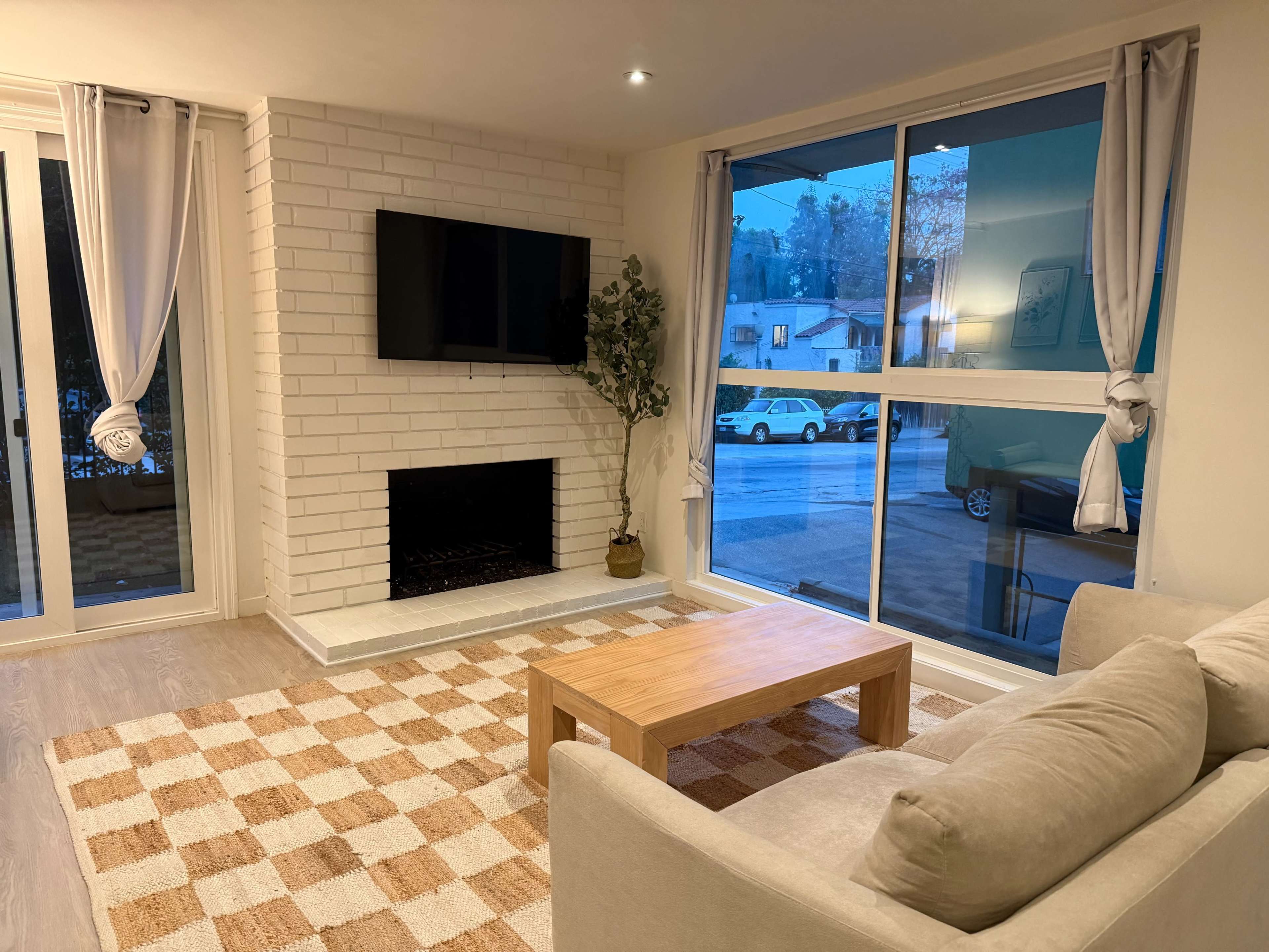 The image shows a living room with a light-colored sofa, a wooden coffee table, a white brick fireplace, and large windows allowing natural light.
