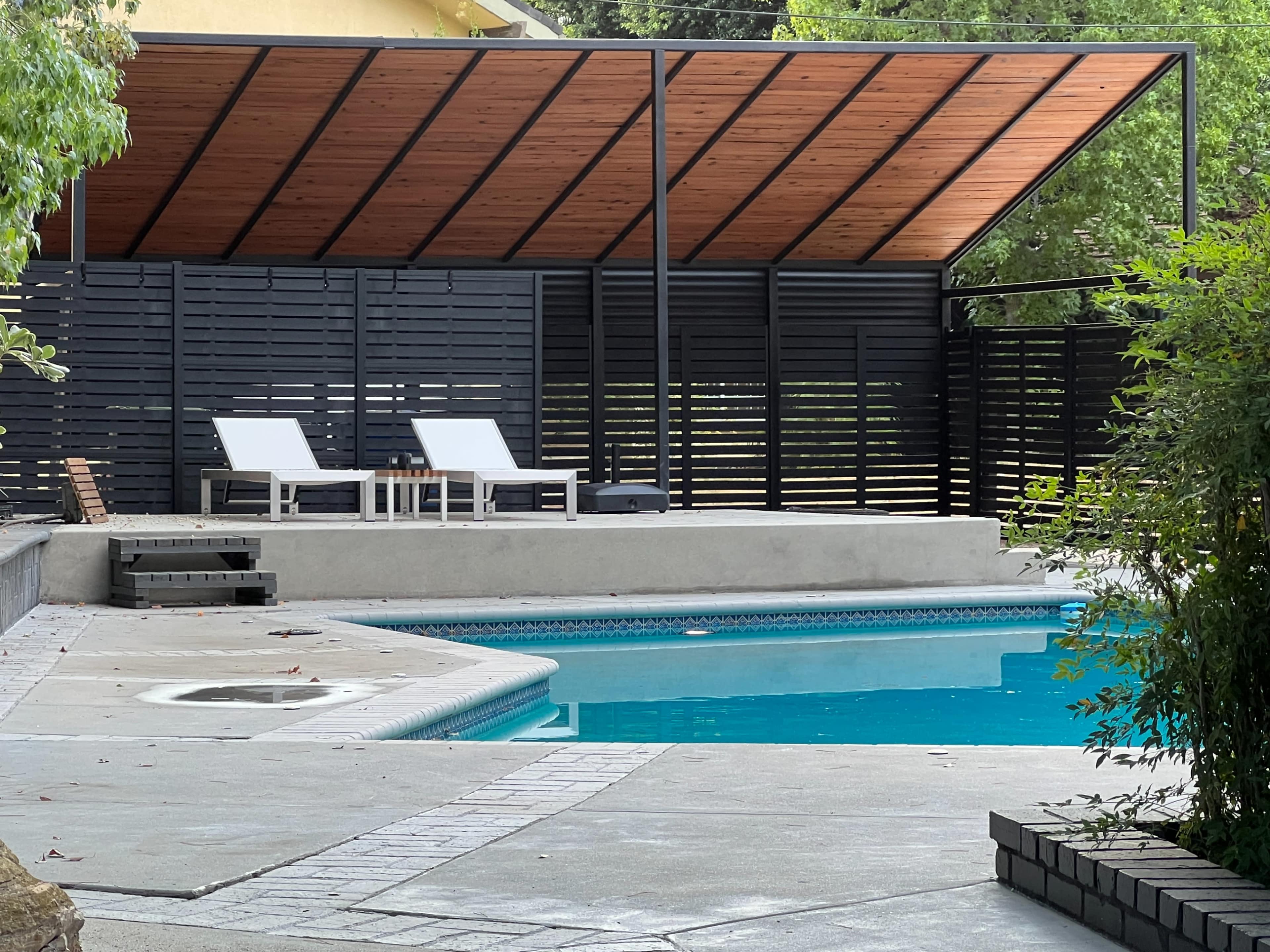 A modern pool area with a covered seating space, two lounge chairs, and a landscaped border.