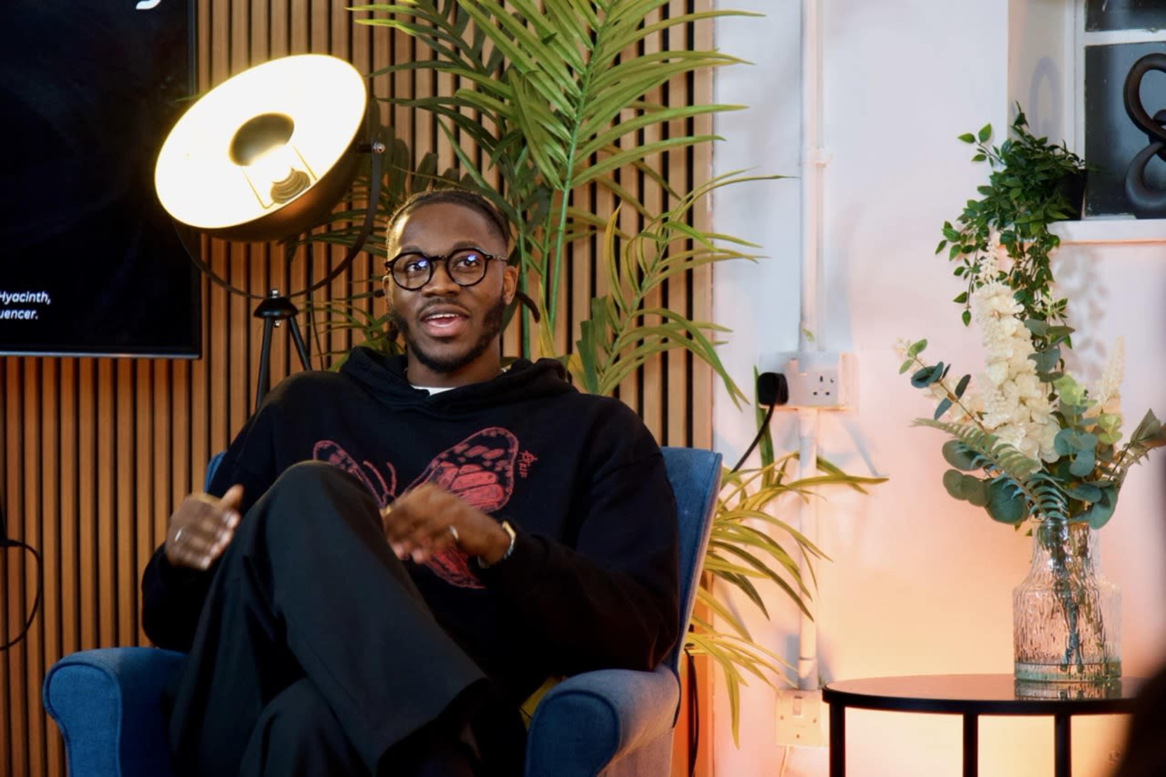 A man wearing glasses and a black hoodie sits in a blue chair, surrounded by plants, with a lamp and a small table decorated with flowers nearby.
