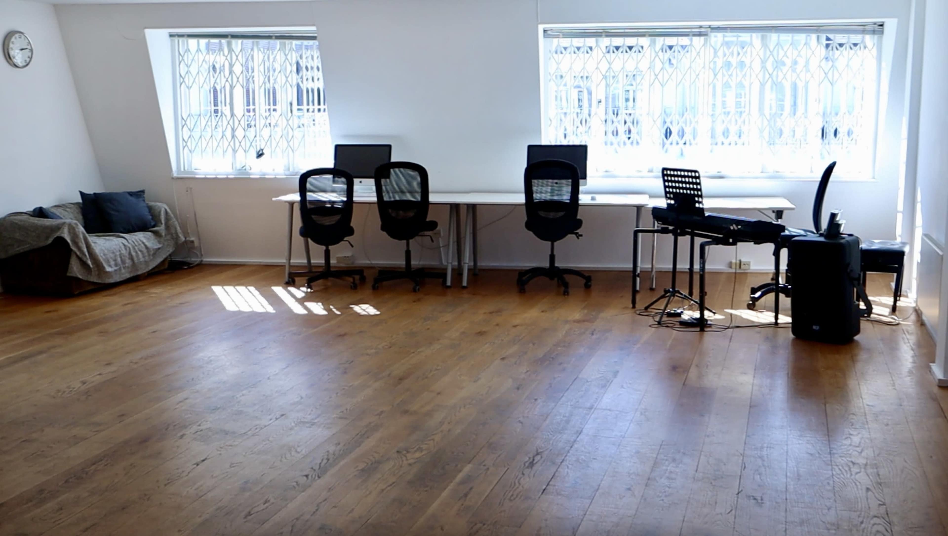 A spacious room with wooden flooring, featuring two desks with chairs on one side and a couch on the other side.