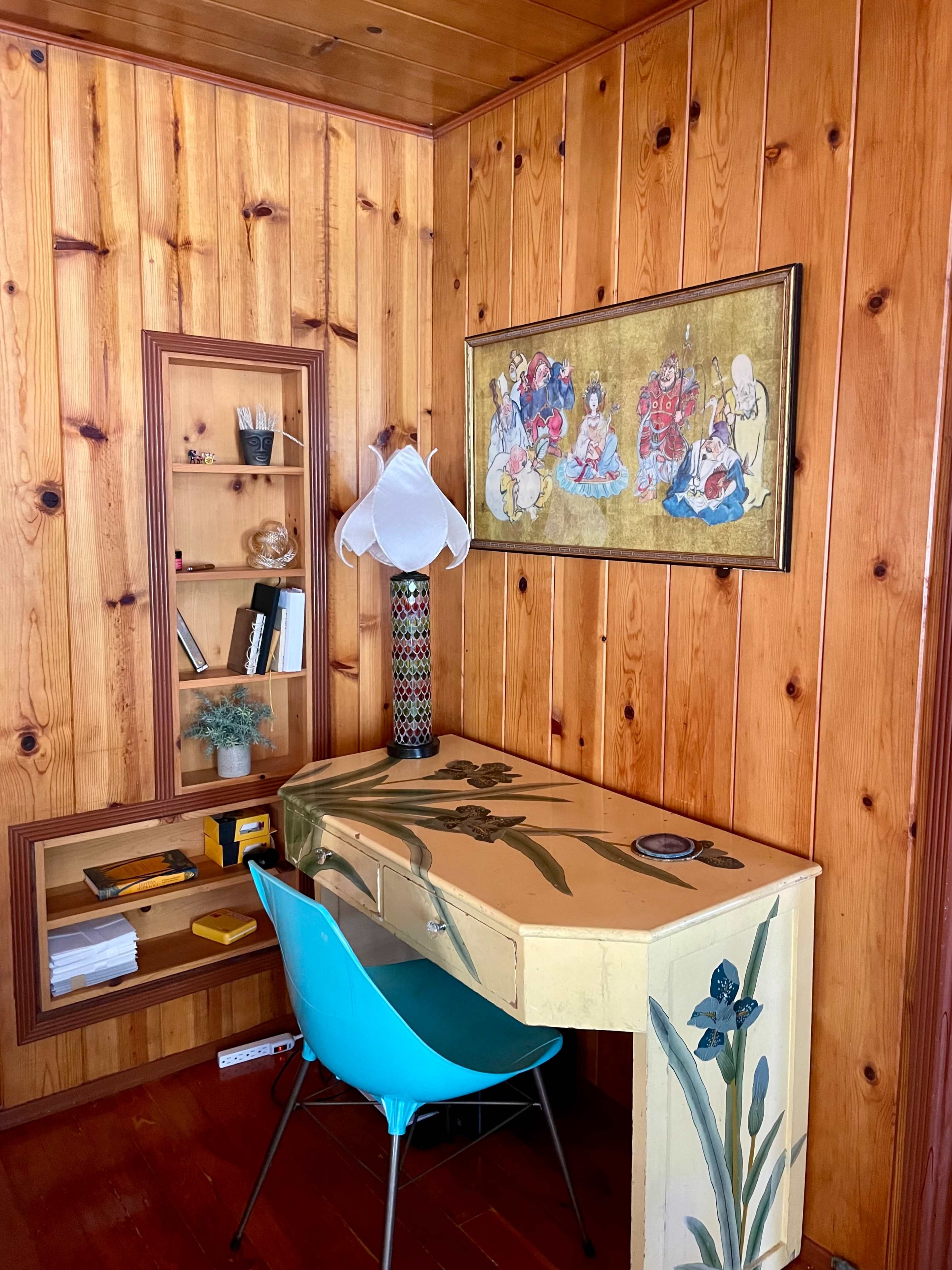 The image shows a small corner workspace with a colorful desk, a blue chair, and shelves displaying books and decorative items against wooden paneled walls.