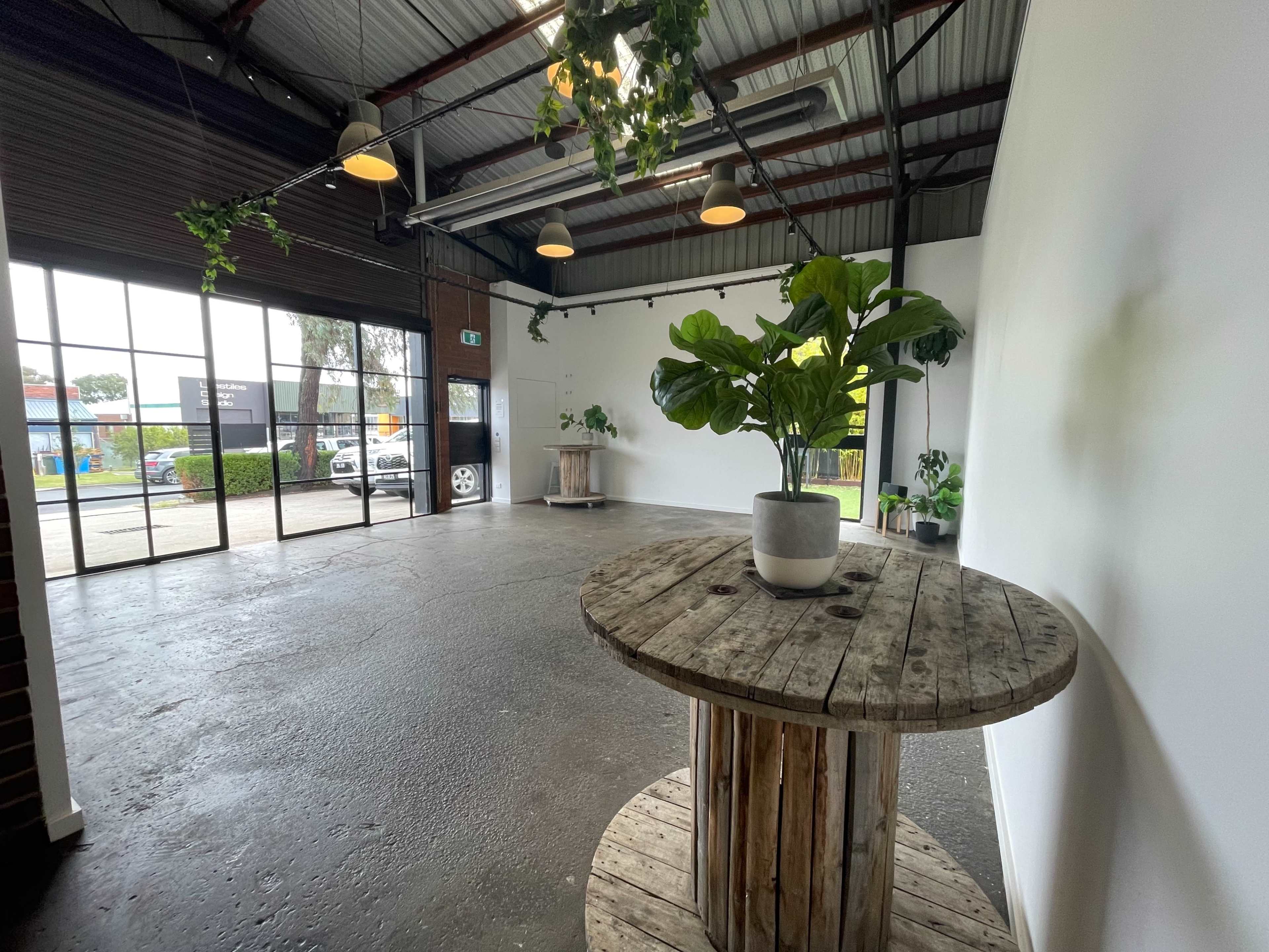 The image shows a spacious, empty room with a wooden table and a potted plant, featuring large windows allowing natural light to enter.