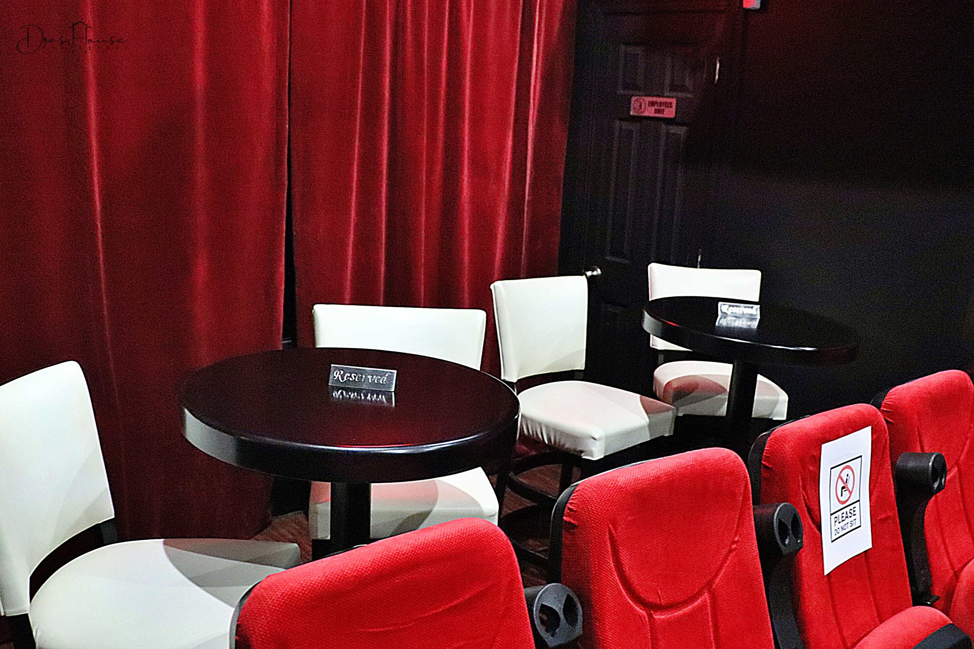 A small seating area with white chairs and round tables is positioned in front of red velvet curtains and the backs of red seats.
