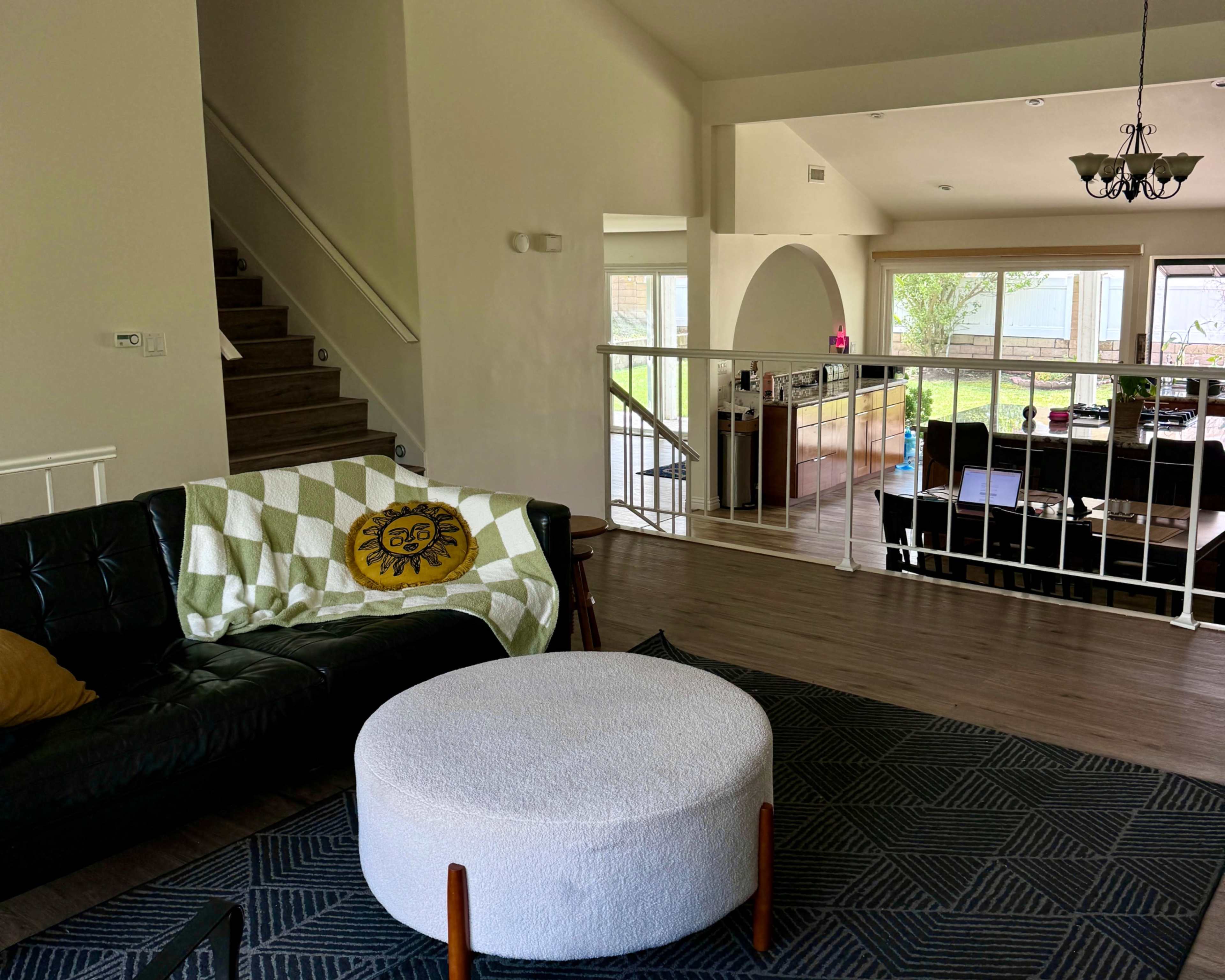 A living room with a black couch, a round white ottoman, and a staircase leading to an upper level, with large windows revealing a backyard.