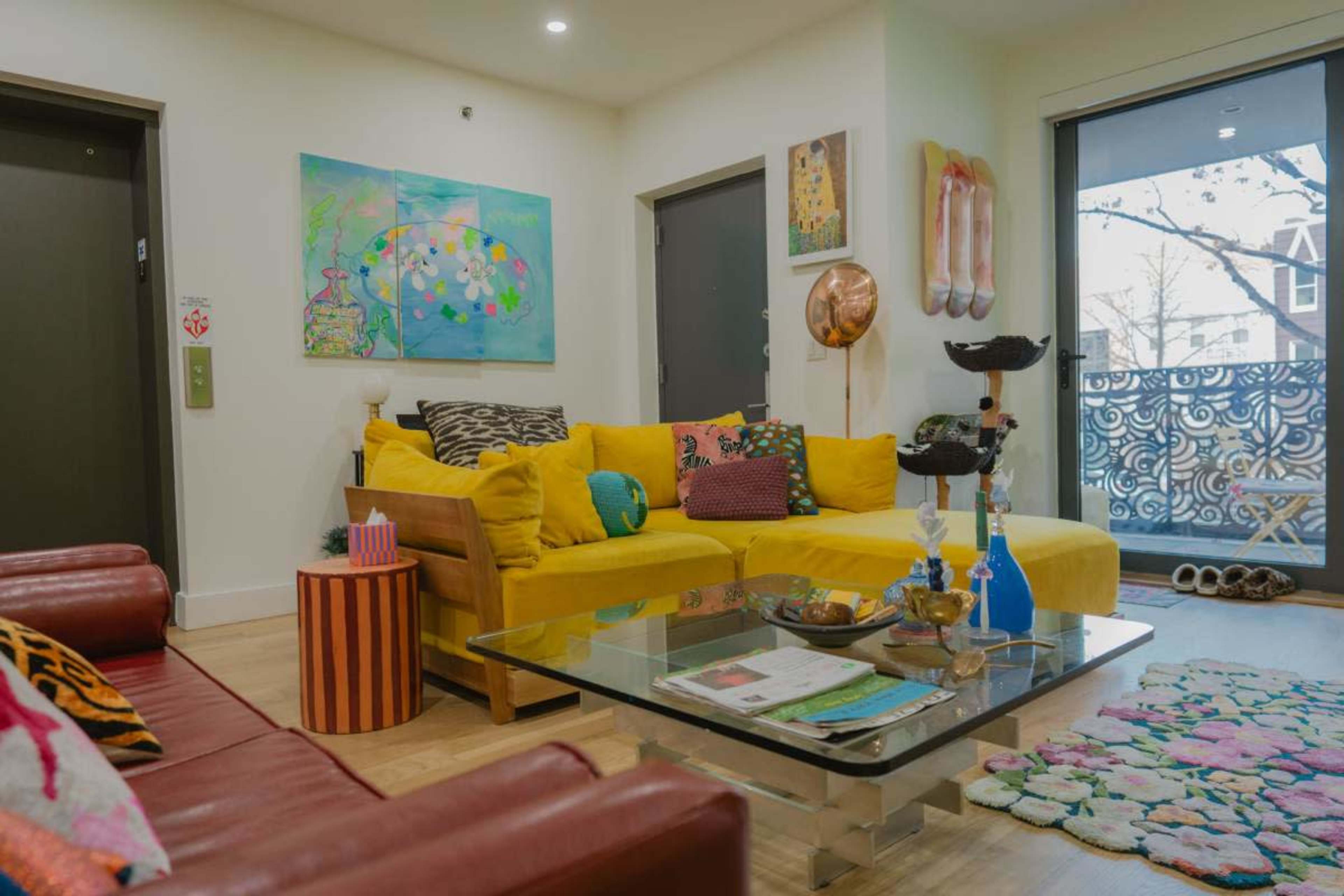 A living room with a yellow sofa, a red leather couch, a coffee table with decorative items, and colorful artwork on the walls.