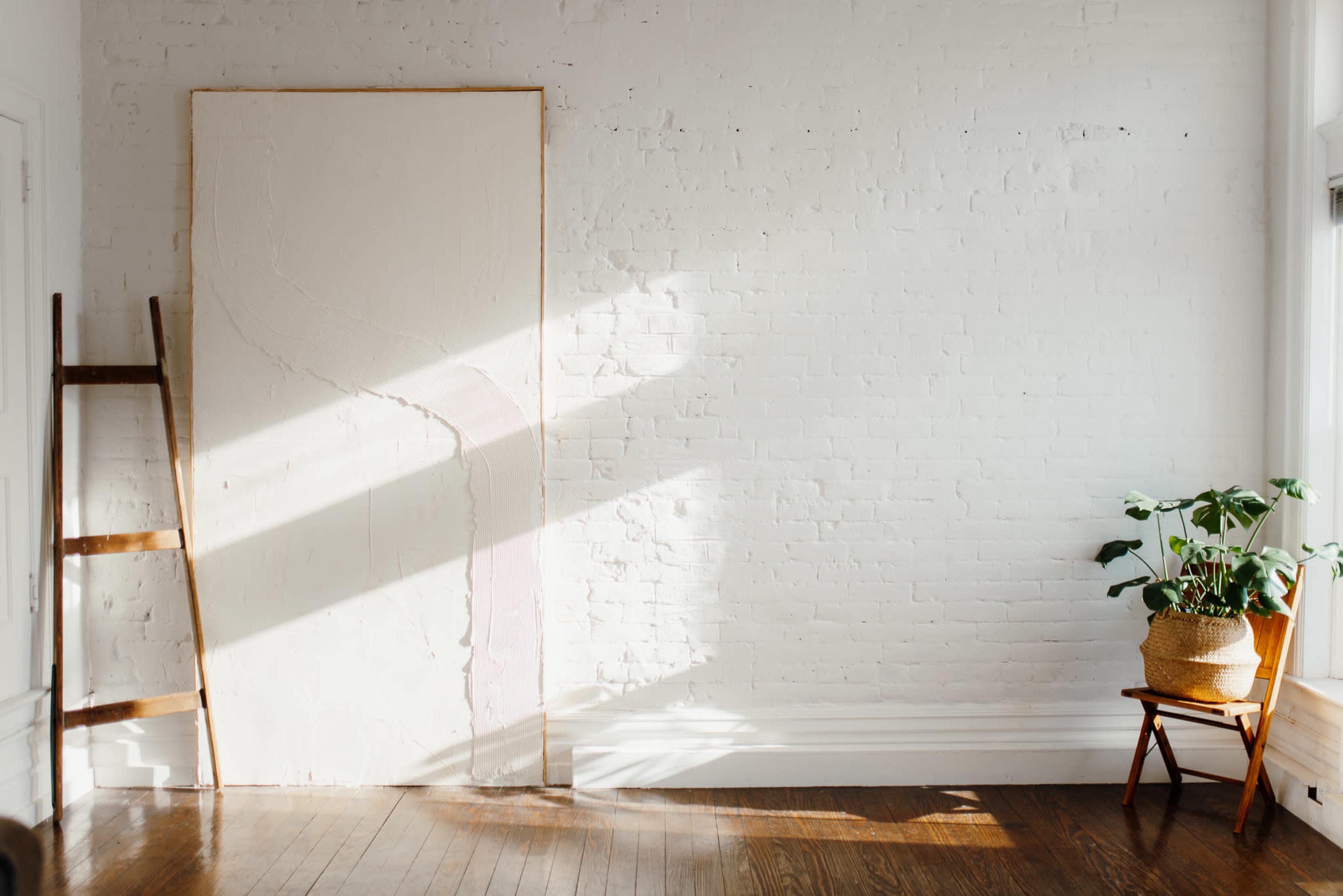 A wooden ladder leans against a white brick wall beside a large, blank canvas, while a potted plant sits next to a wooden chair in a sunlit room.