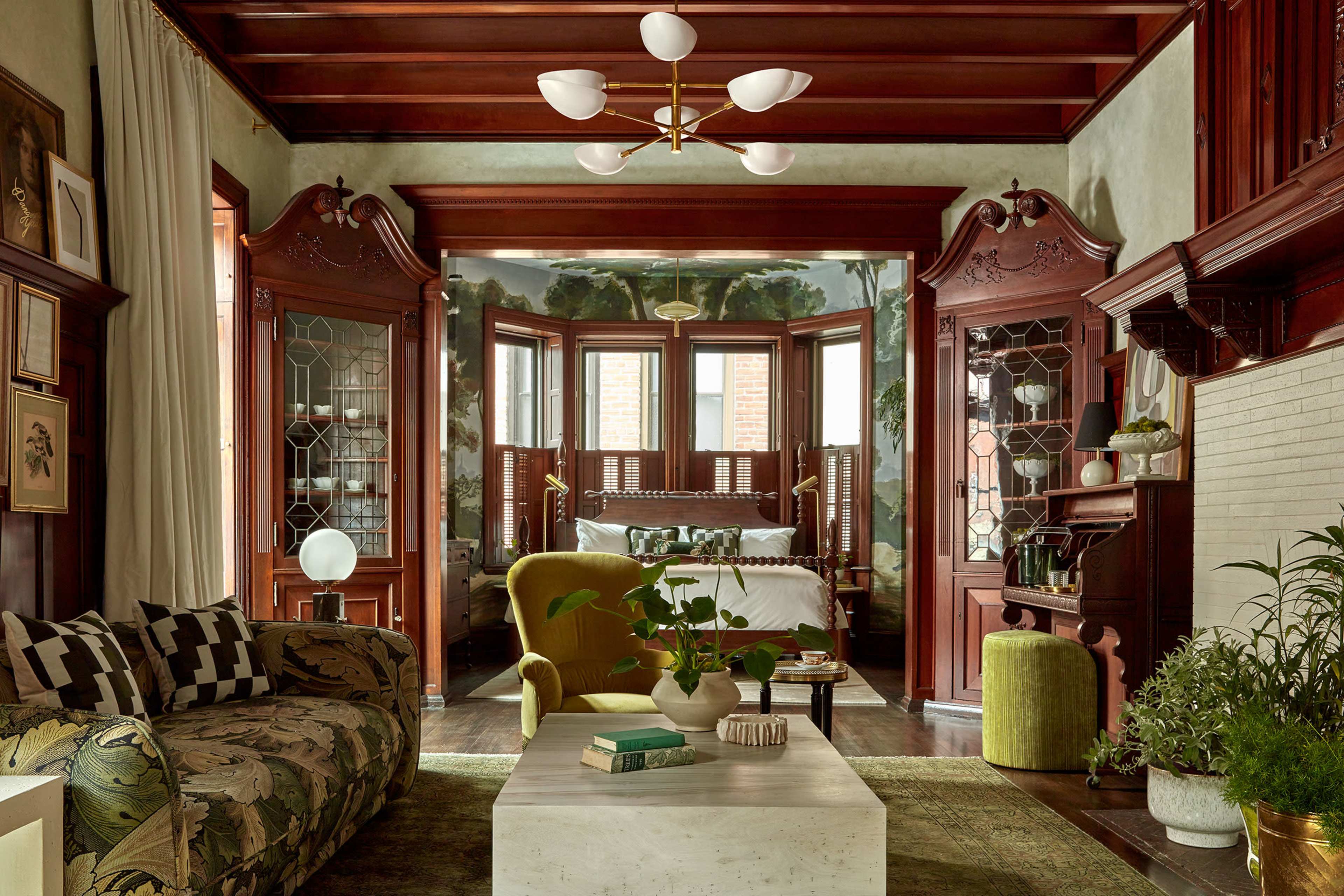 The image shows a spacious, elegantly designed living room featuring wooden beams, large windows, and a mix of contemporary and vintage furniture, including a patterned sofa and a green accent chair.