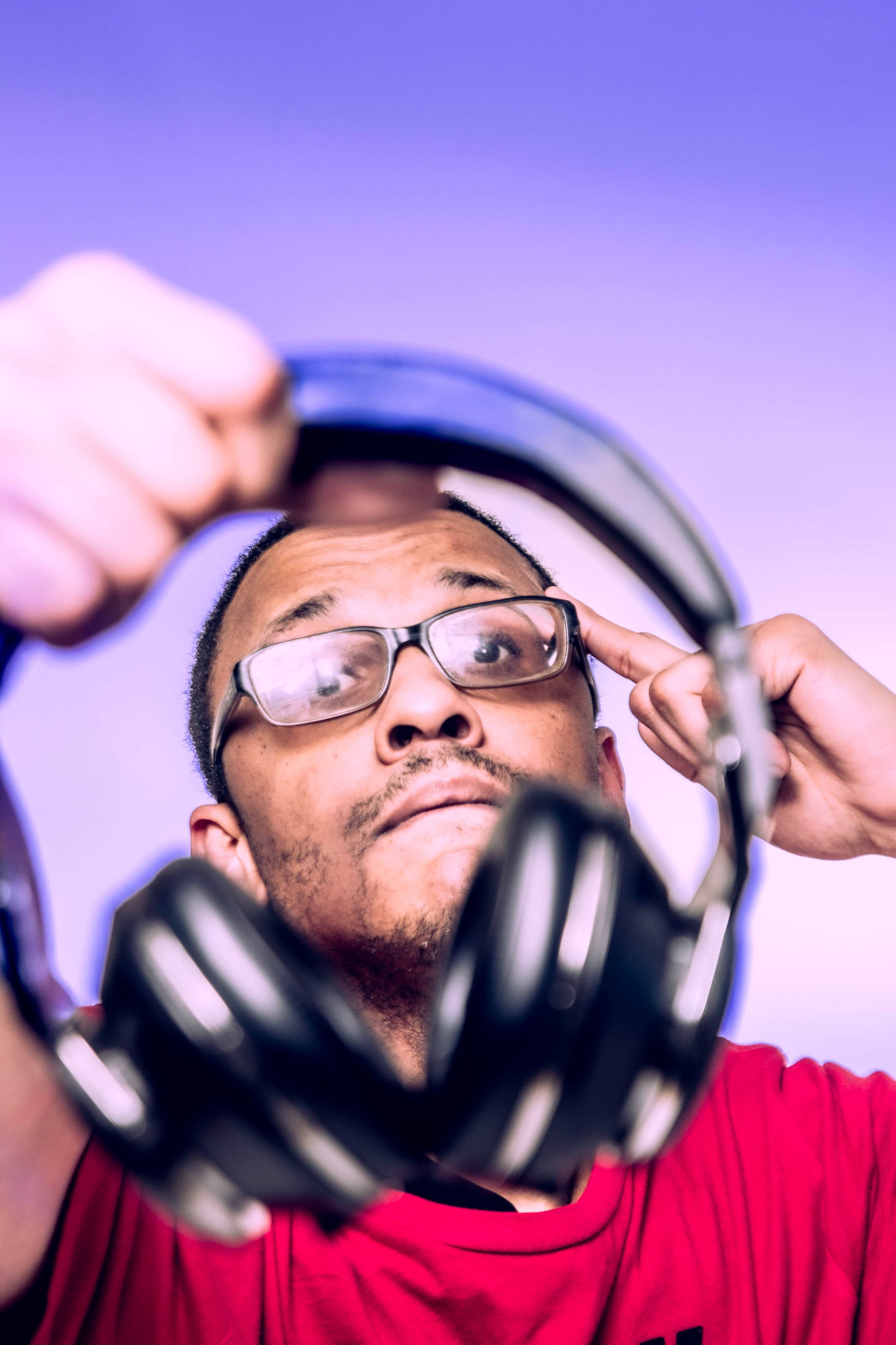 A person wearing glasses holds a pair of headphones in front of their face against a purple background.
