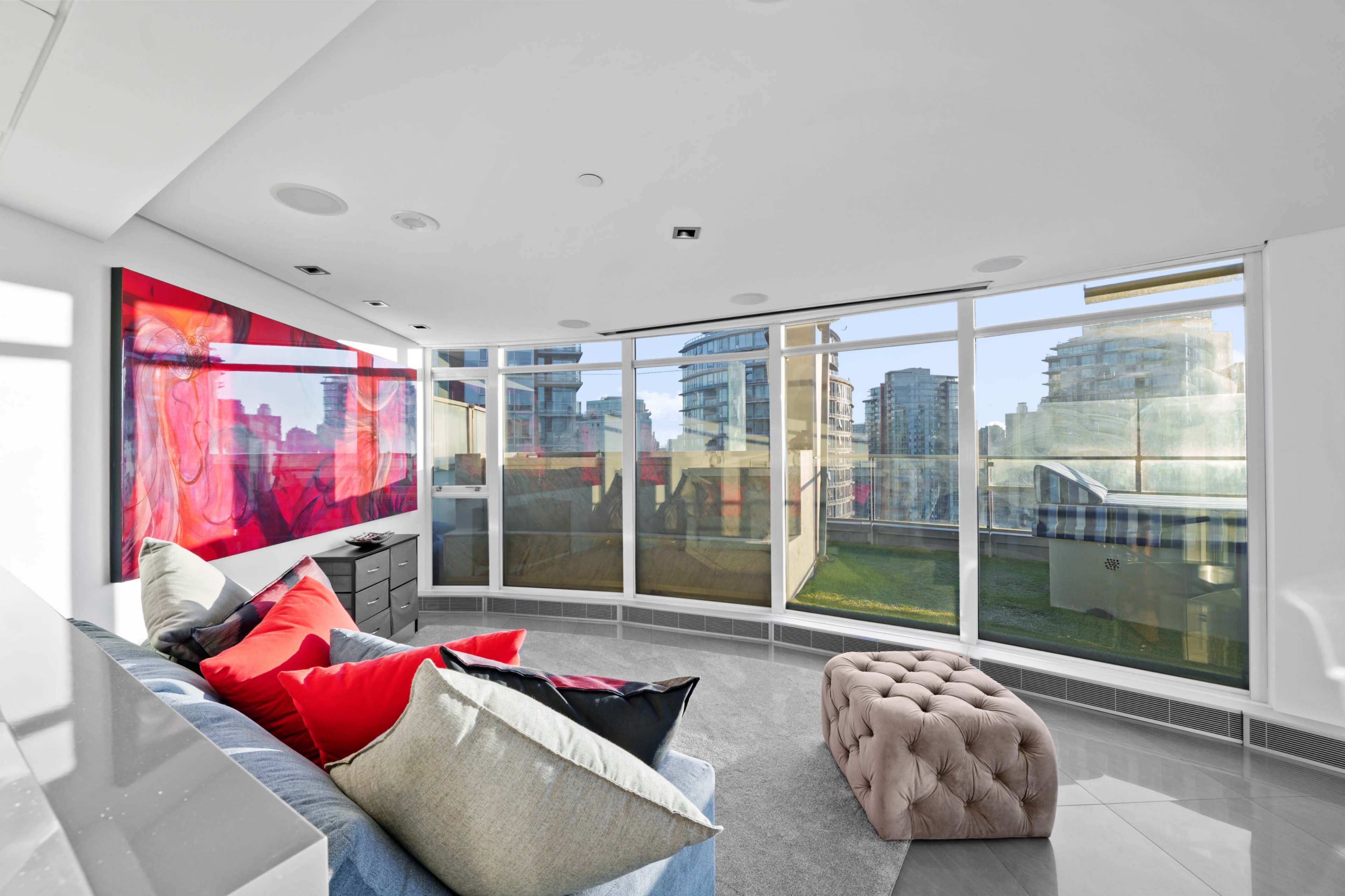 The image shows a modern living space with large windows, a colorful wall art piece, and a plush seating area featuring decorative pillows and an ottoman.