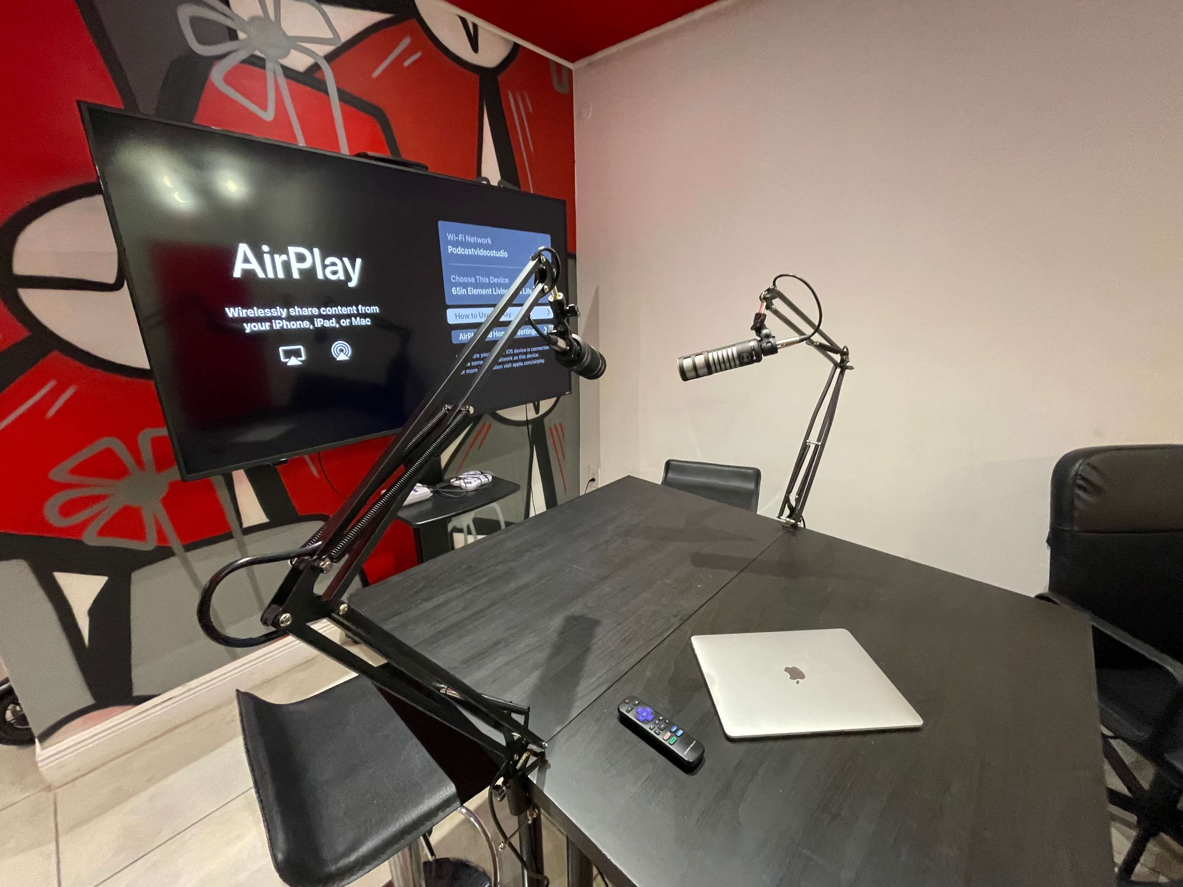 A black table with a laptop and remote is positioned under a wall-mounted screen displaying an "AirPlay" interface, alongside a microphone setup on adjustable arms.