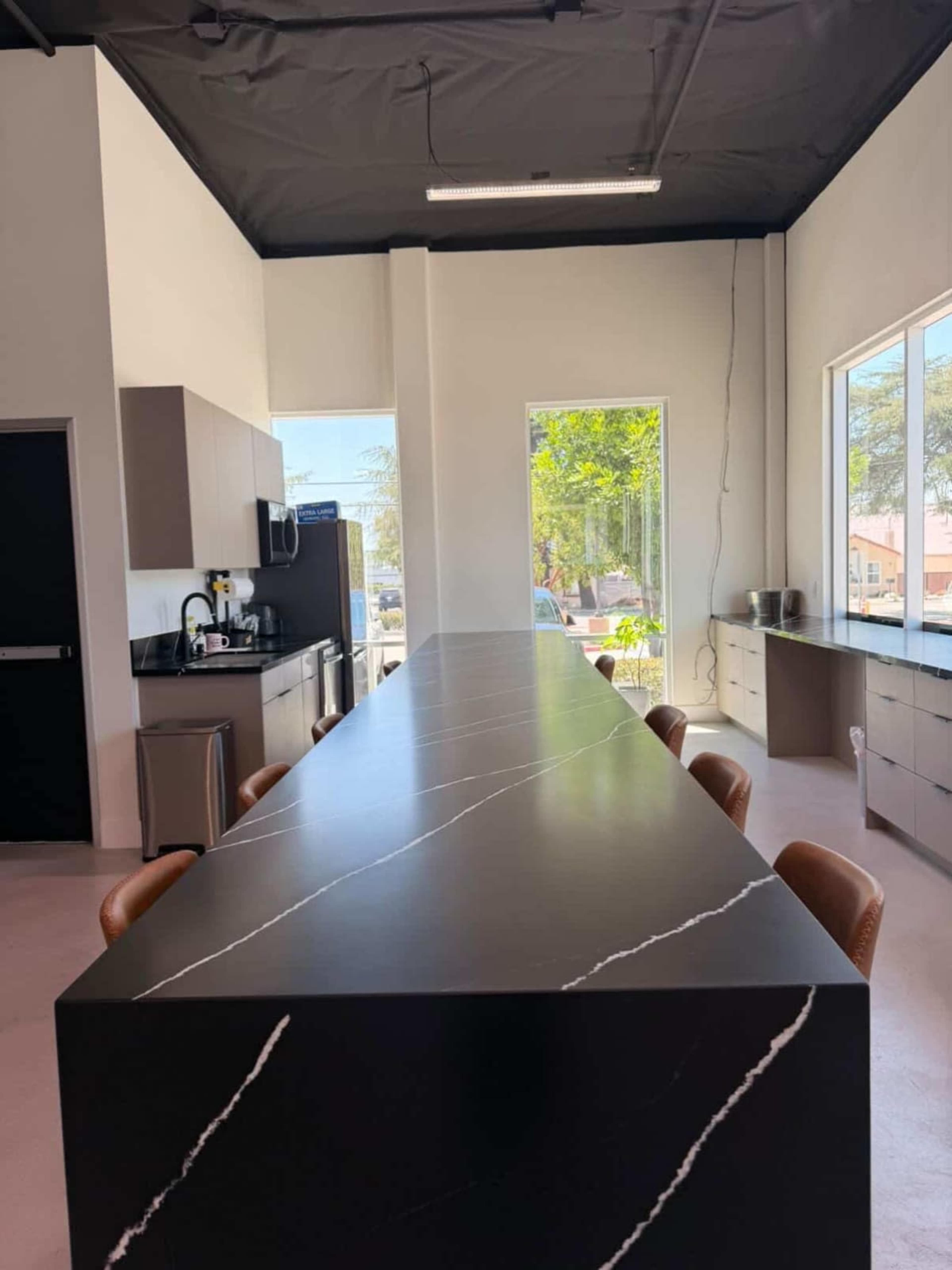 The image shows a modern kitchen with a long black marble countertop and brown leather chairs, featuring large windows that allow natural light to enter.