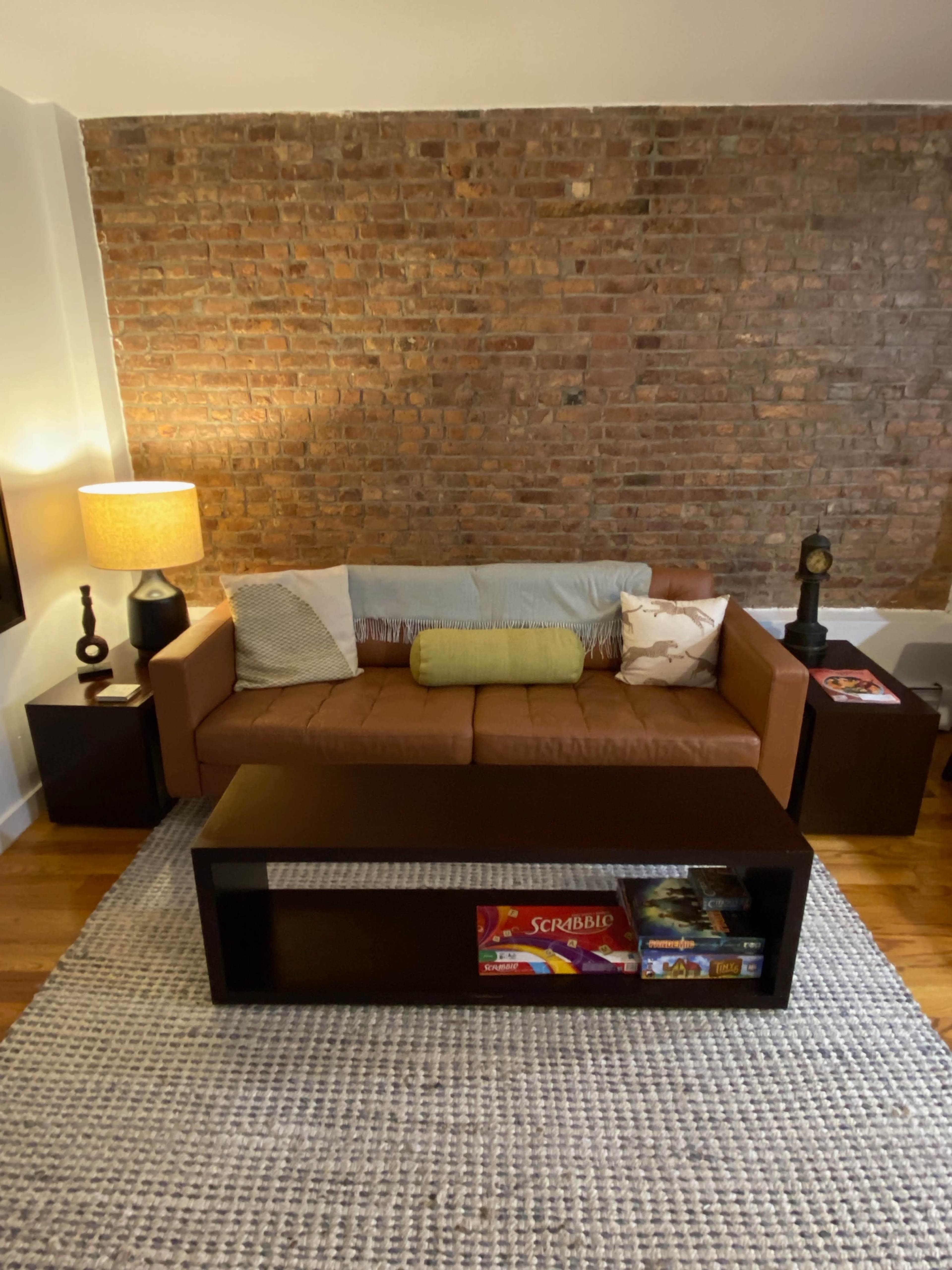 A living room with a brown sofa against a brick wall, a coffee table displaying a board game, and a lamp on a side table.