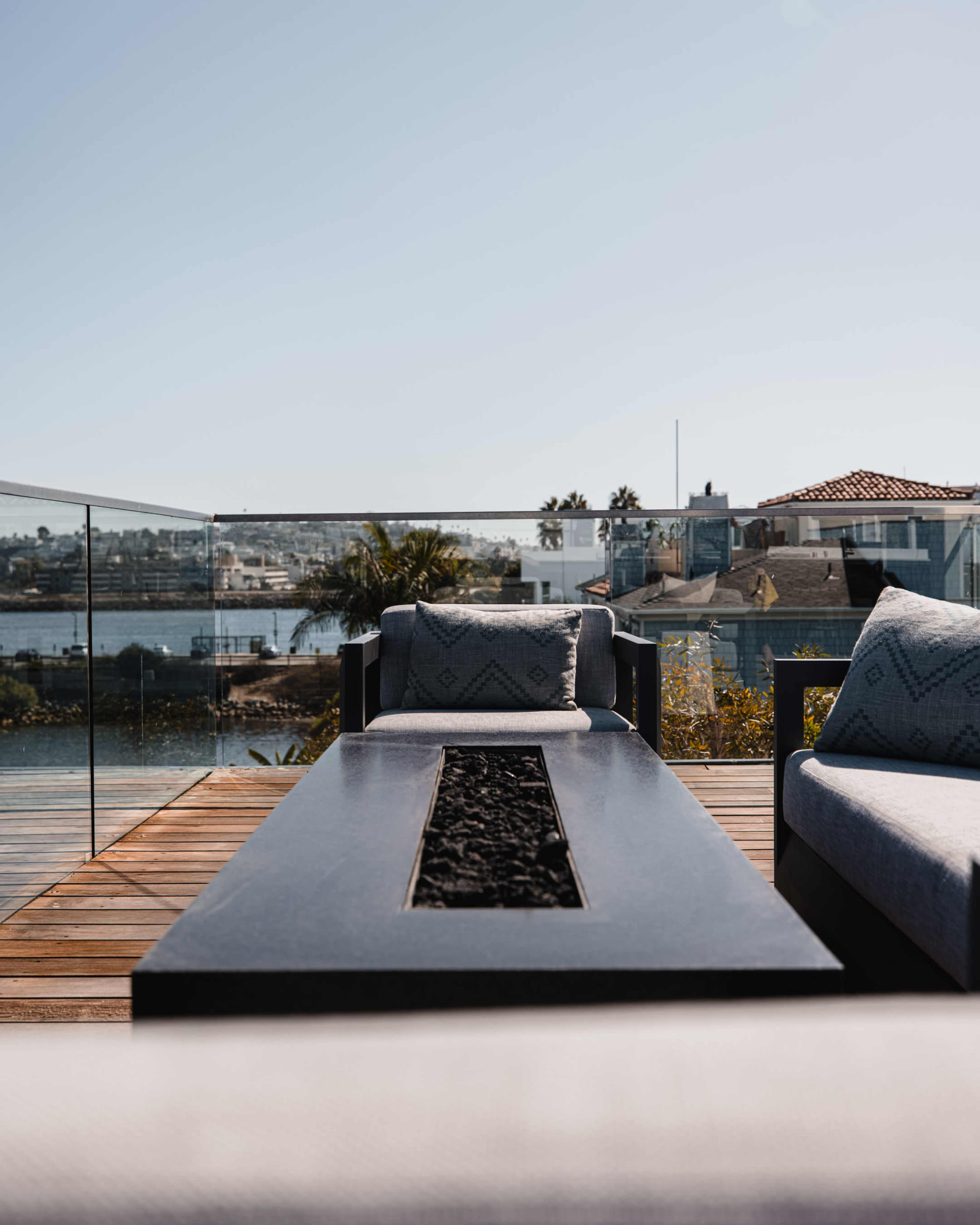 A modern outdoor seating area features a sleek table with a fire pit, surrounded by cushions and a view of the water and nearby homes.