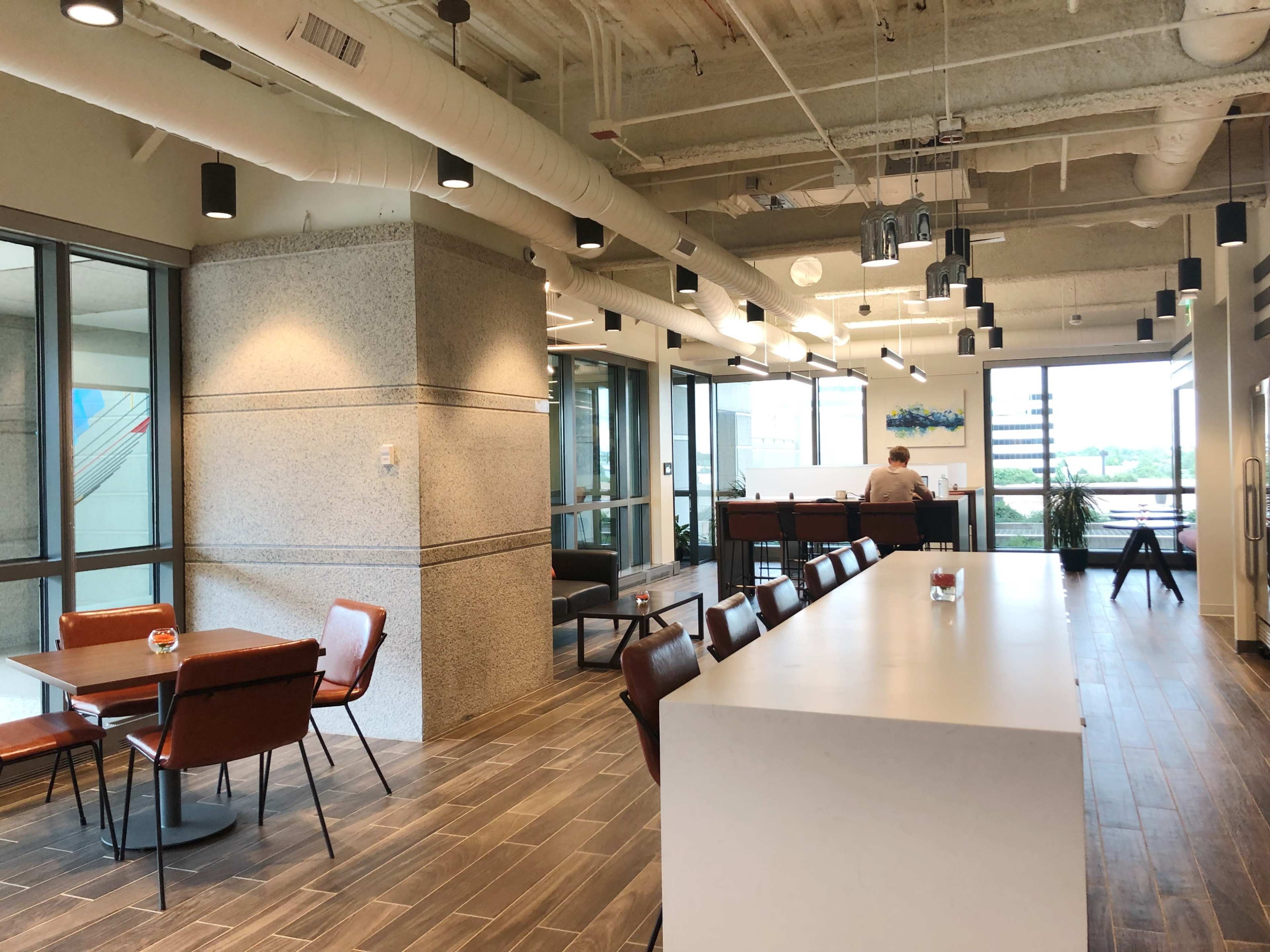 A modern office space with a dining area featuring tables and chairs on one side, and a bar-style seating area along a long counter on the other, with large windows allowing natural light.