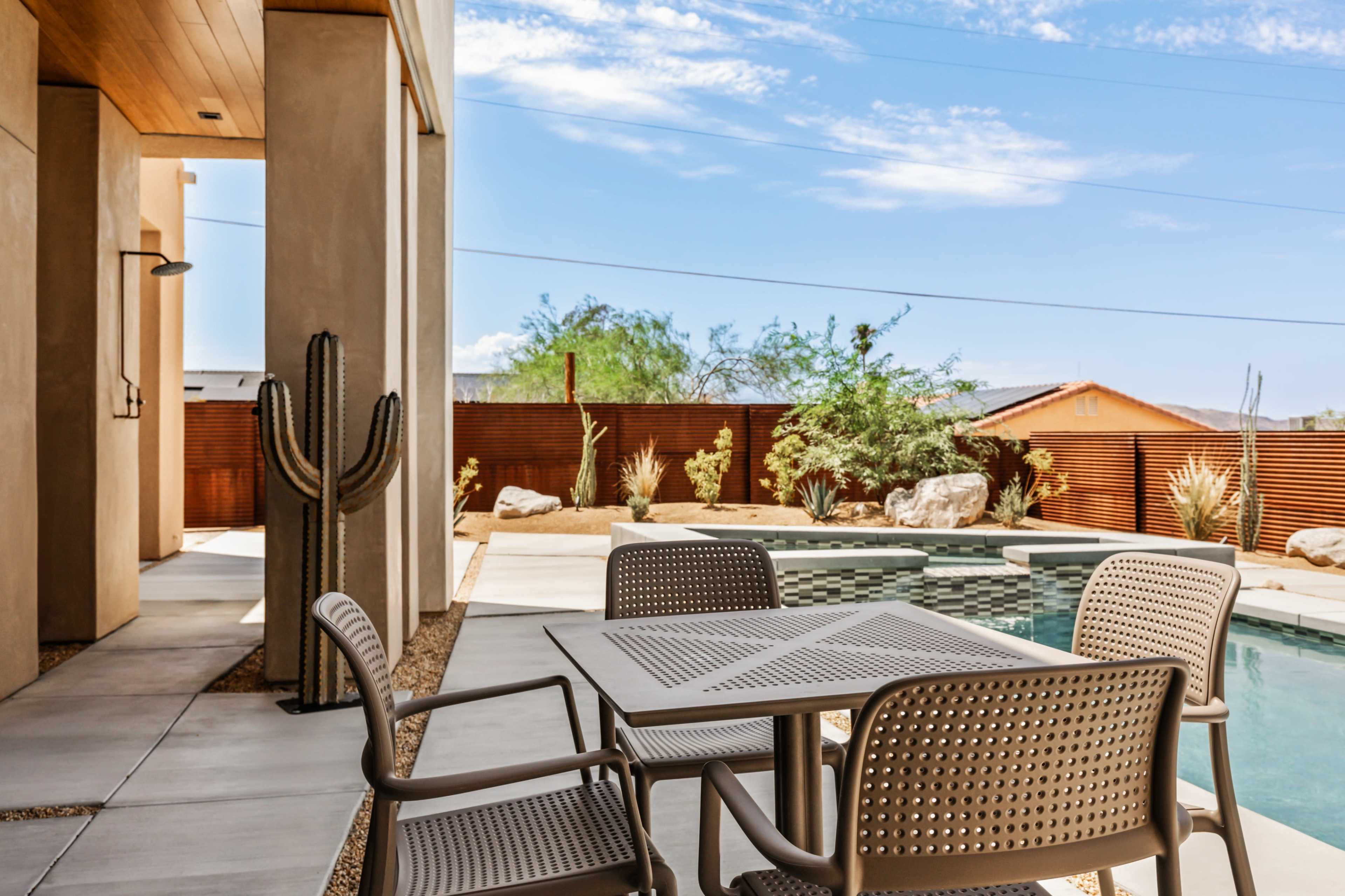 A modern patio features a table and chairs overlooking a swimming pool, with desert landscaping and a cactus nearby.