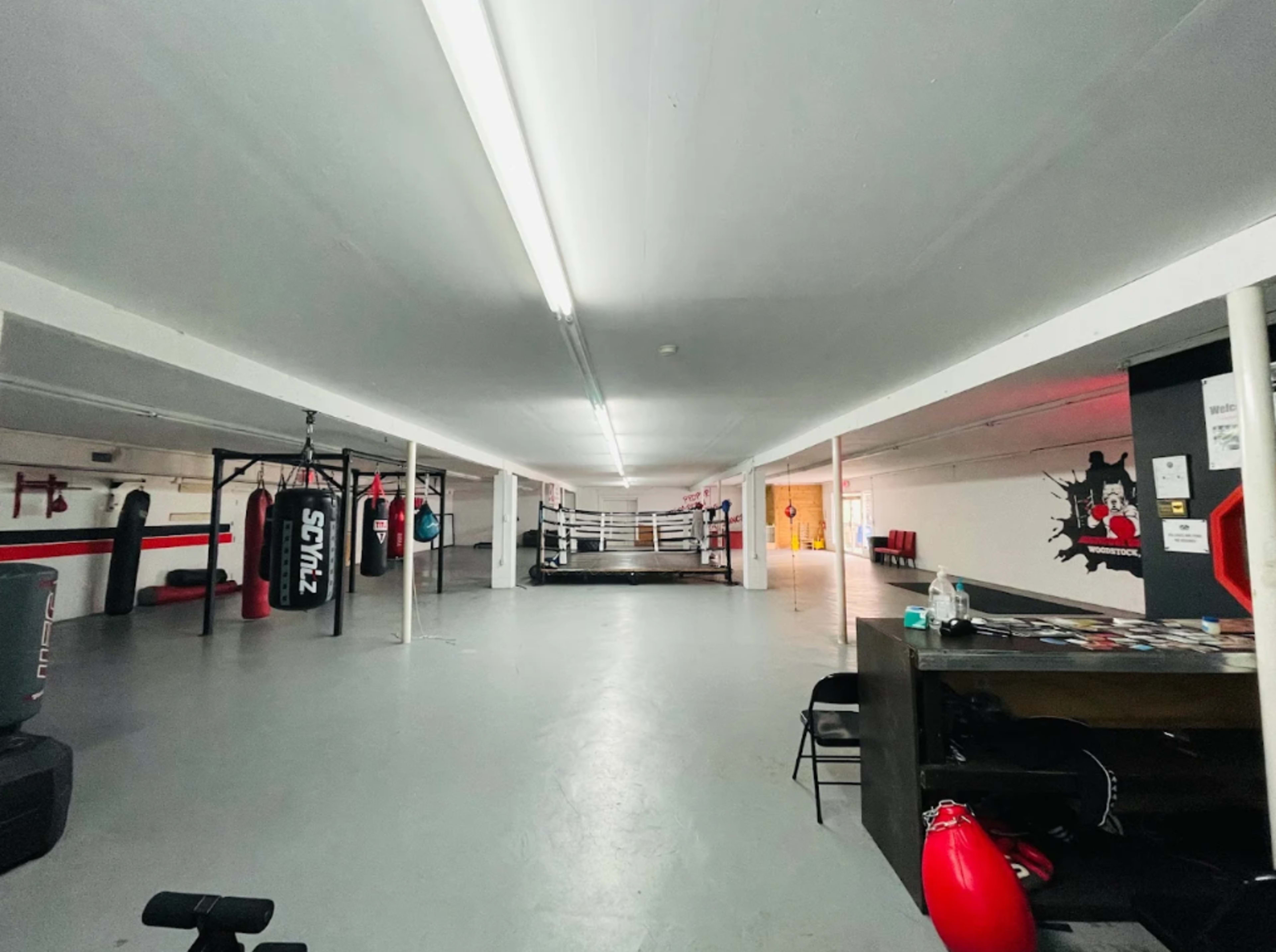 The image shows a spacious gym interior with punching bags, a boxing ring, and a seating area.