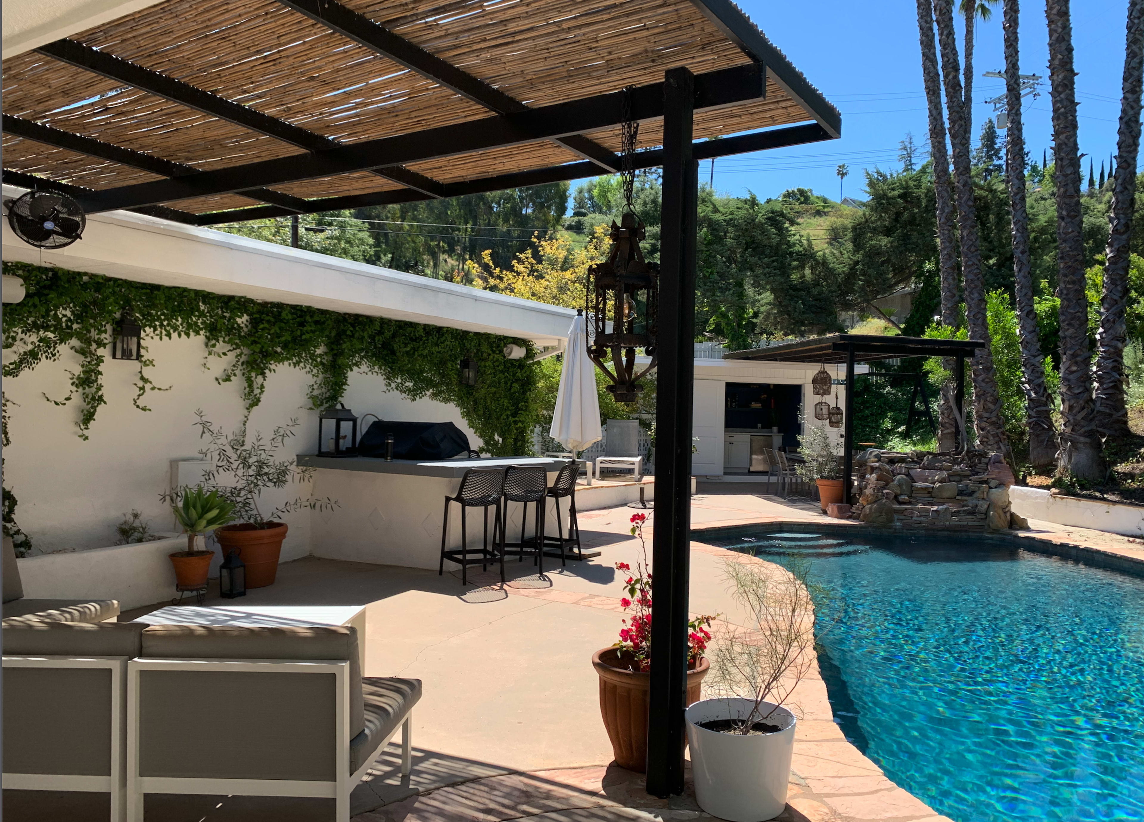 The image shows a backyard patio with seating, a swimming pool, and a barbecue area surrounded by greenery and palm trees.