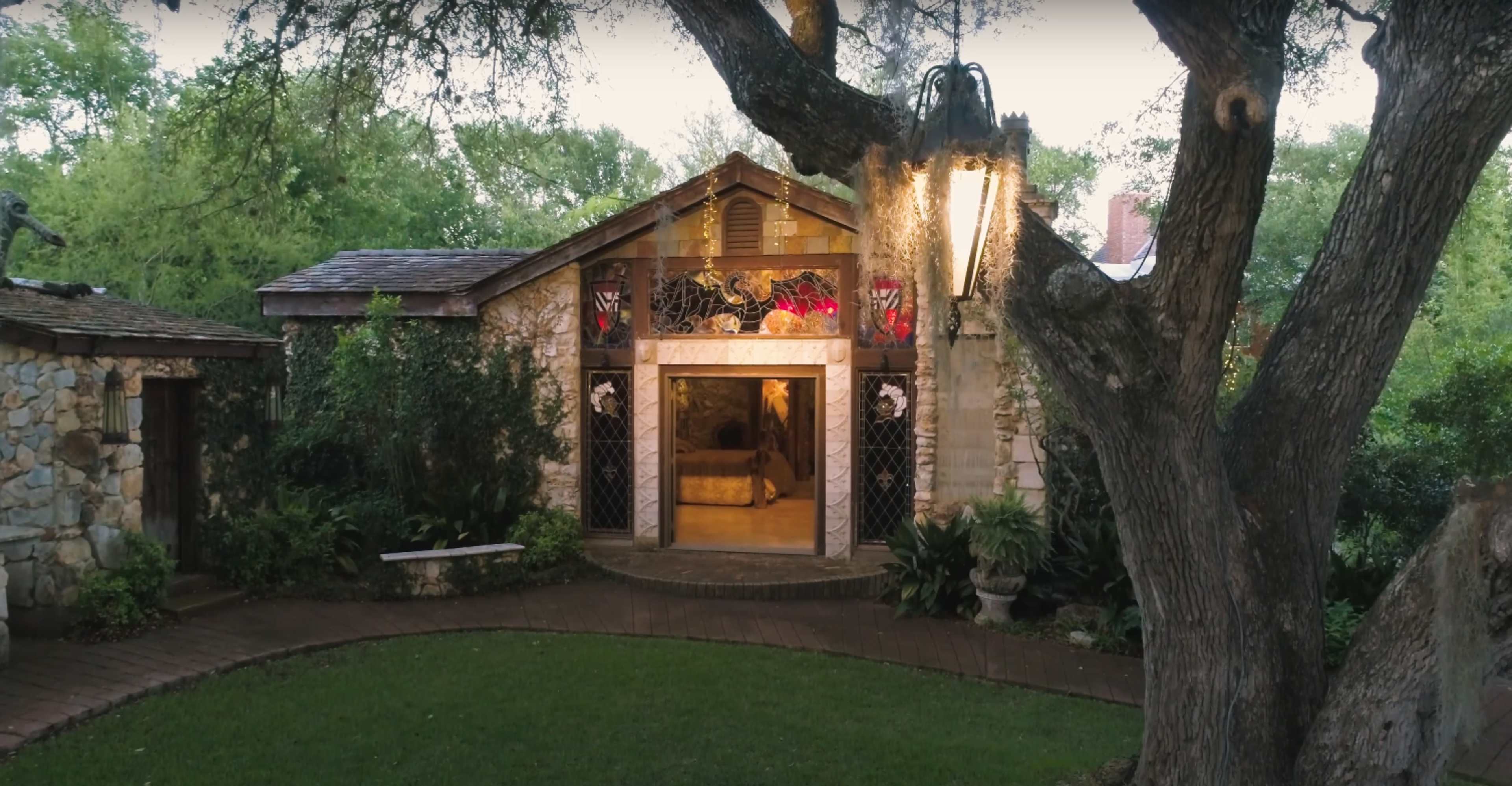 A stone cottage with a wooden front door is surrounded by lush greenery and a large tree, illuminated by a lantern.
