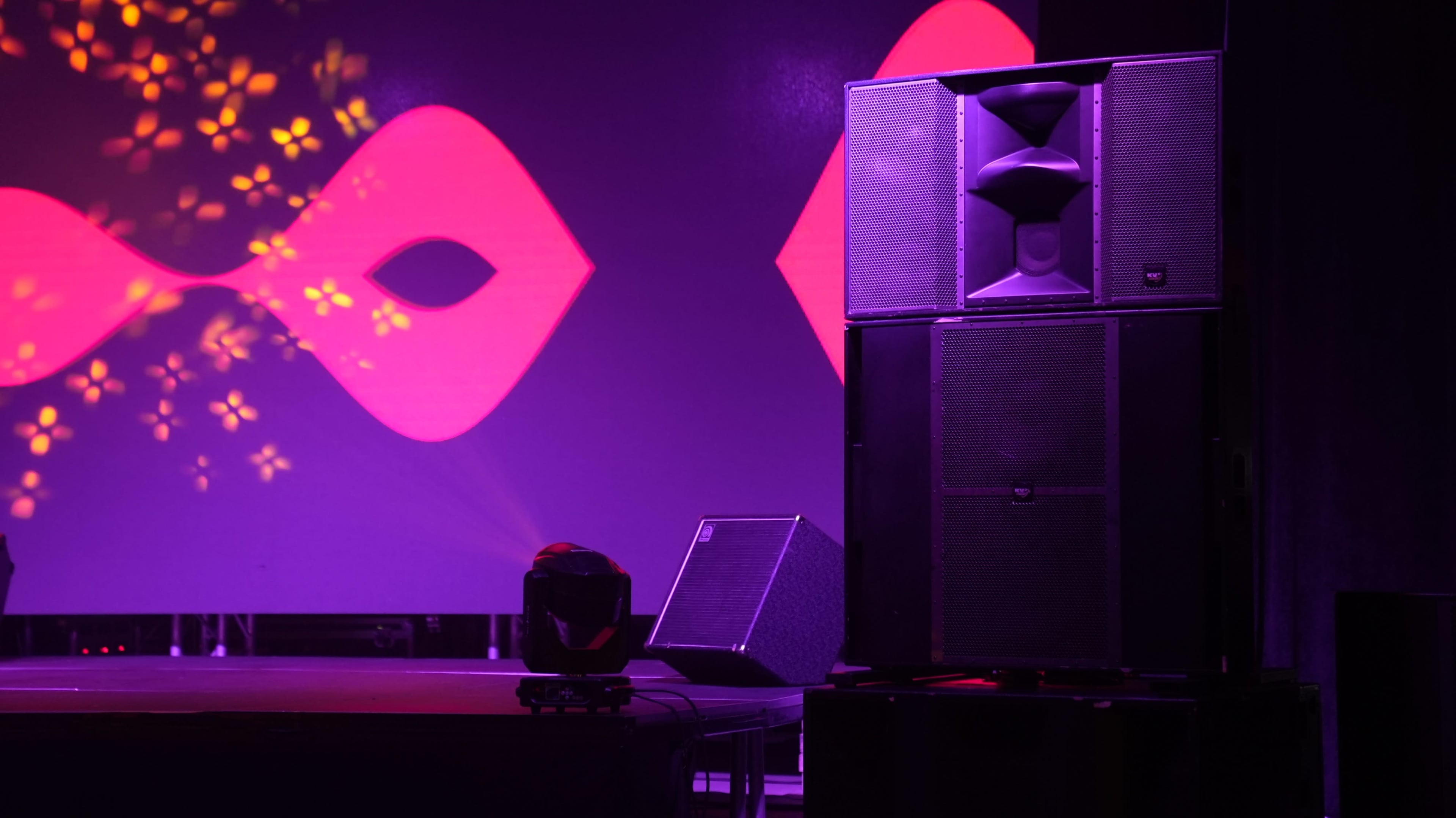 A large sound system is positioned on a stage in front of a colorful backdrop featuring abstract shapes and lighting effects.