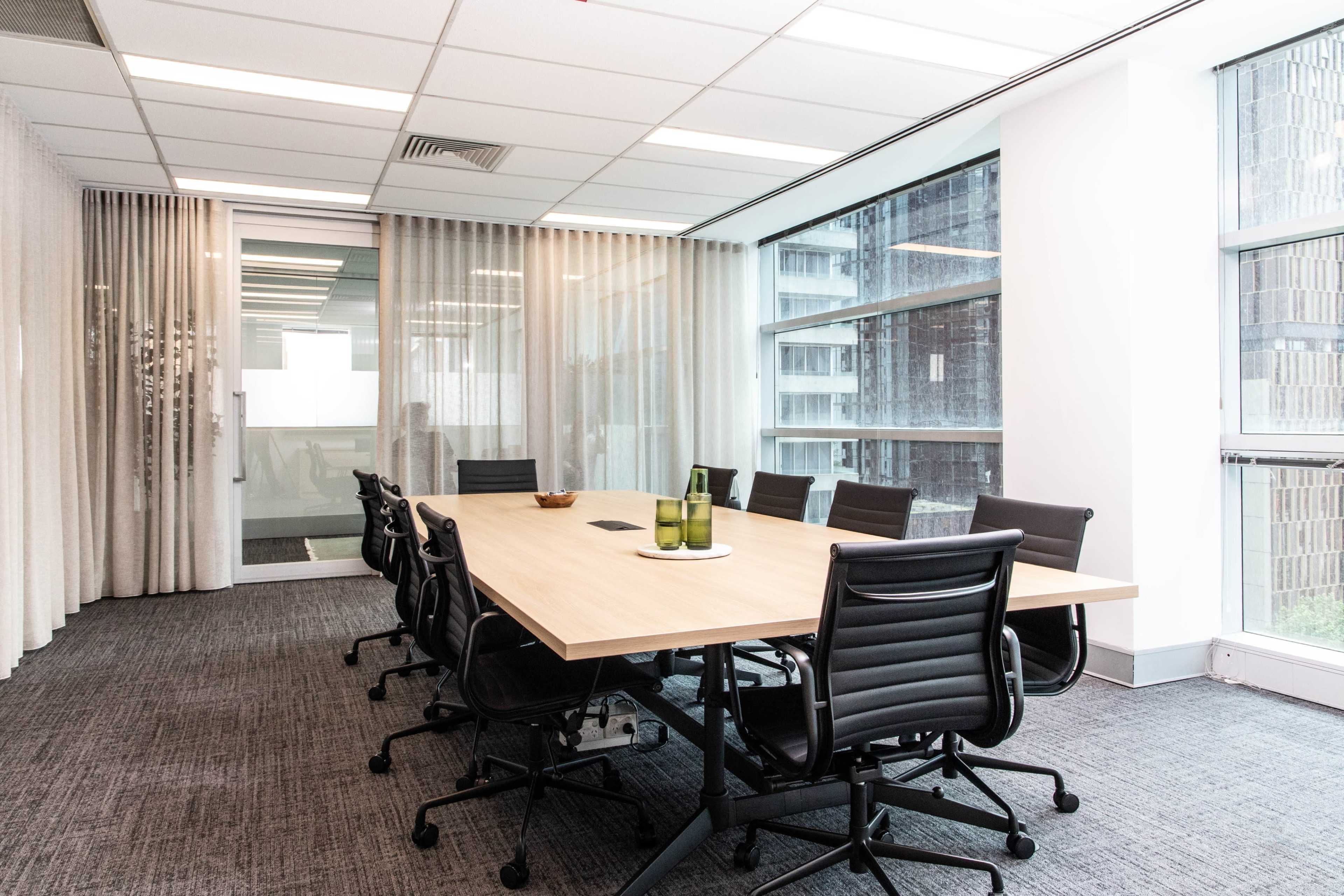 A modern conference room features a long wooden table surrounded by black office chairs, with large windows allowing natural light to enter.